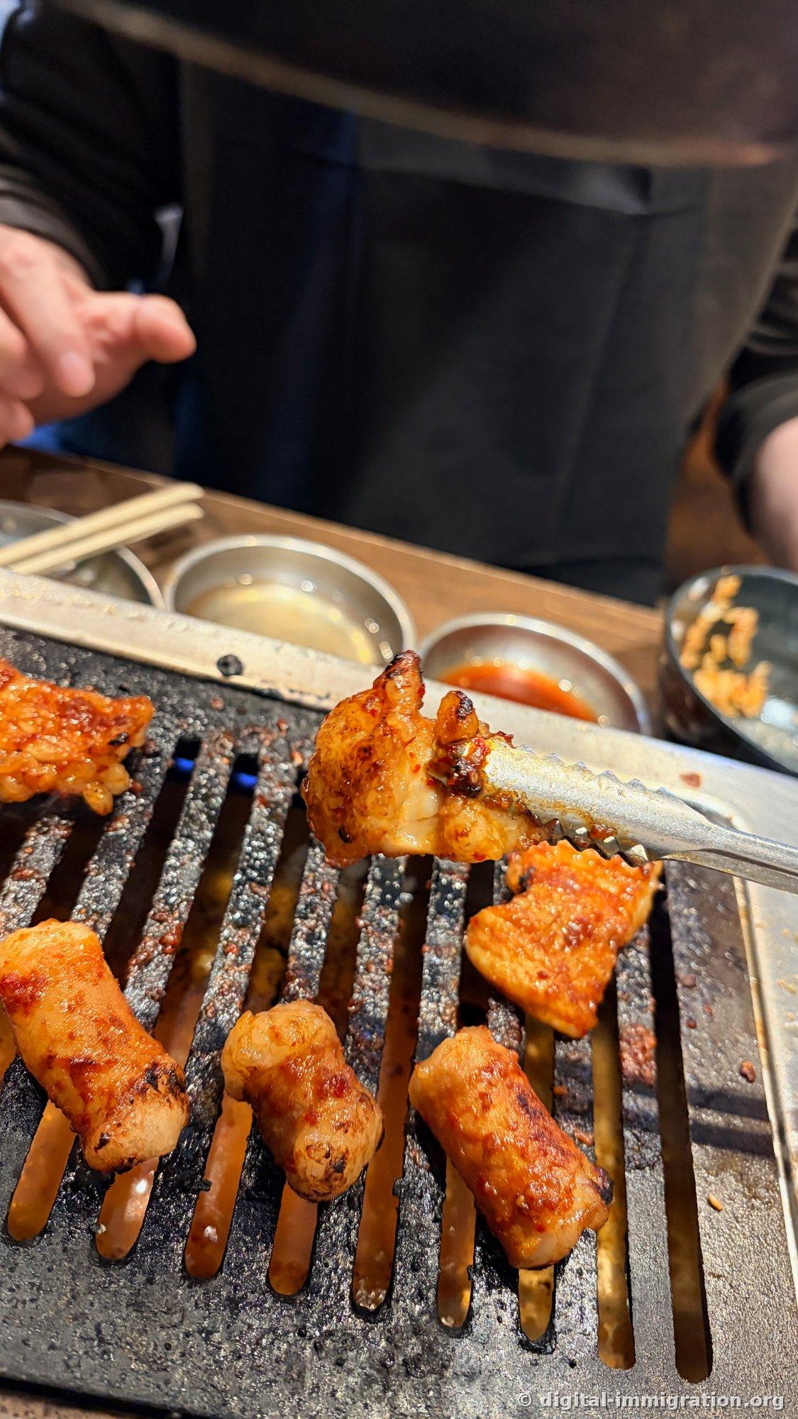 Multiple plump pieces of red-sauced hormon cooking on the grill, one being lifted with tongs; sauce dishes visible in the background