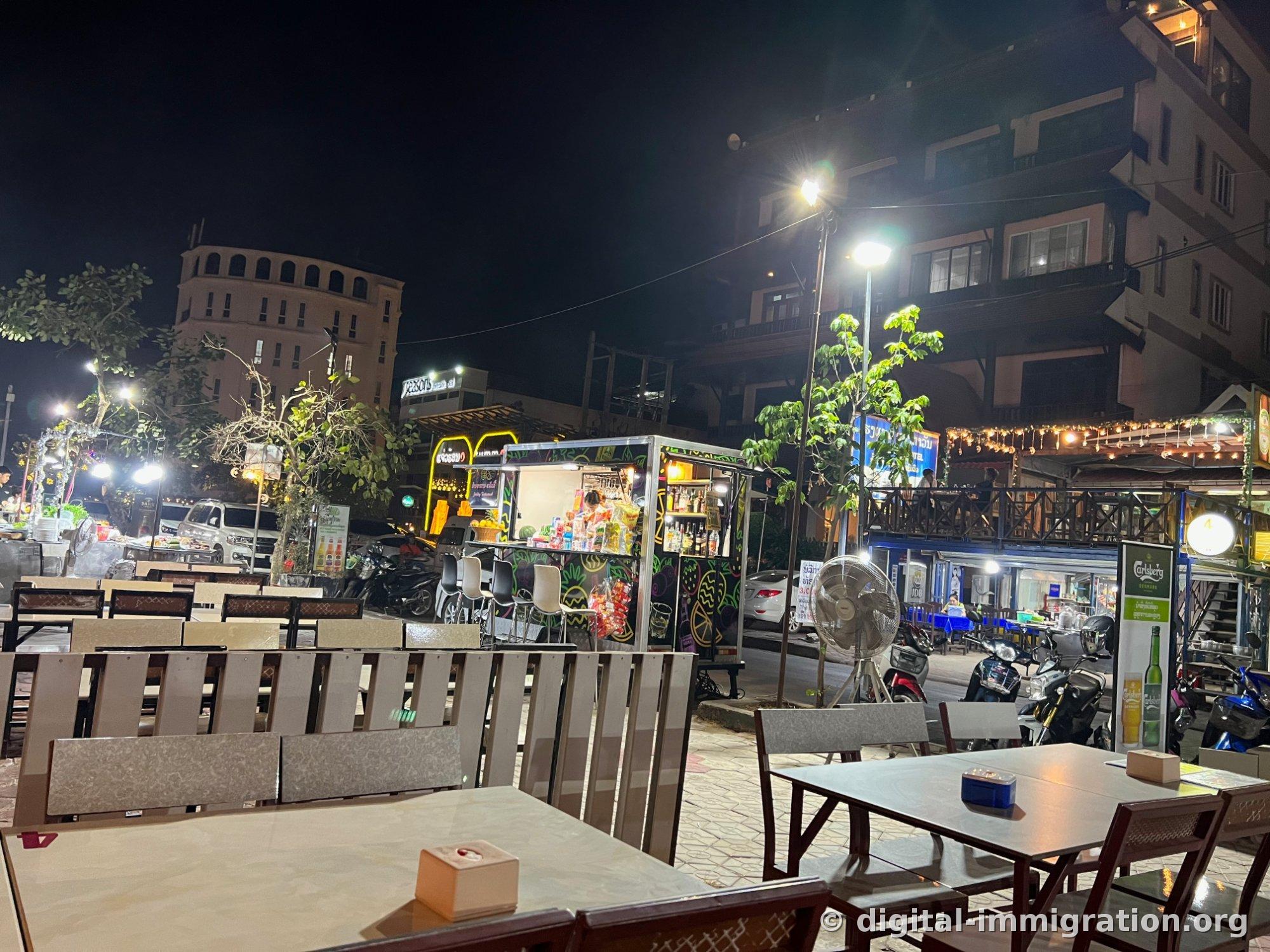 没有客人的露天餐桌后面，街对面有画着荧光涂鸦的流动餐车和Carlsberg广告牌，还有一台大型电风扇的夜间街景