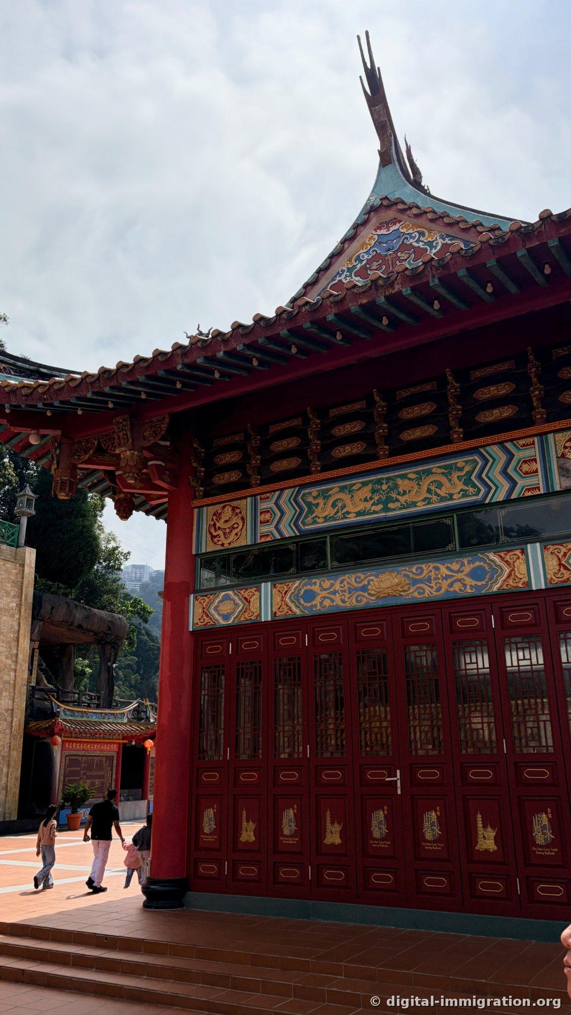 Façade du bâtiment principal du temple avec des colonnes rouges et une peinture murale ornée, les portes en bois pourpre portant le nom du temple et le motif de la pagode en feuille d'or