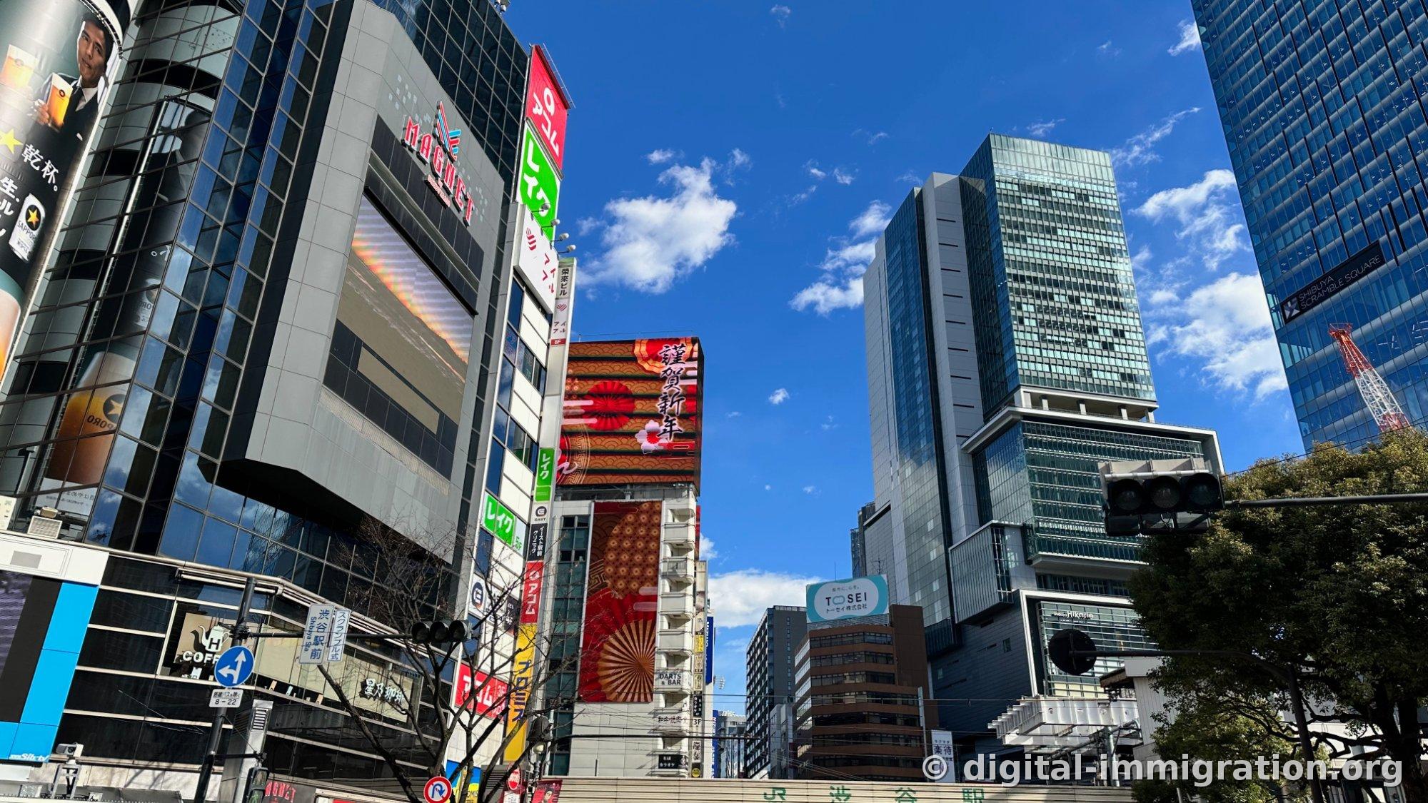Sous le ciel bleu, les bâtiments autour du carrefour Scramble de Shibuya — l'écran MAGNET, l'affiche rouge 謹賀新年, et les bâtiments Scramble Square et Hikarie sont visibles