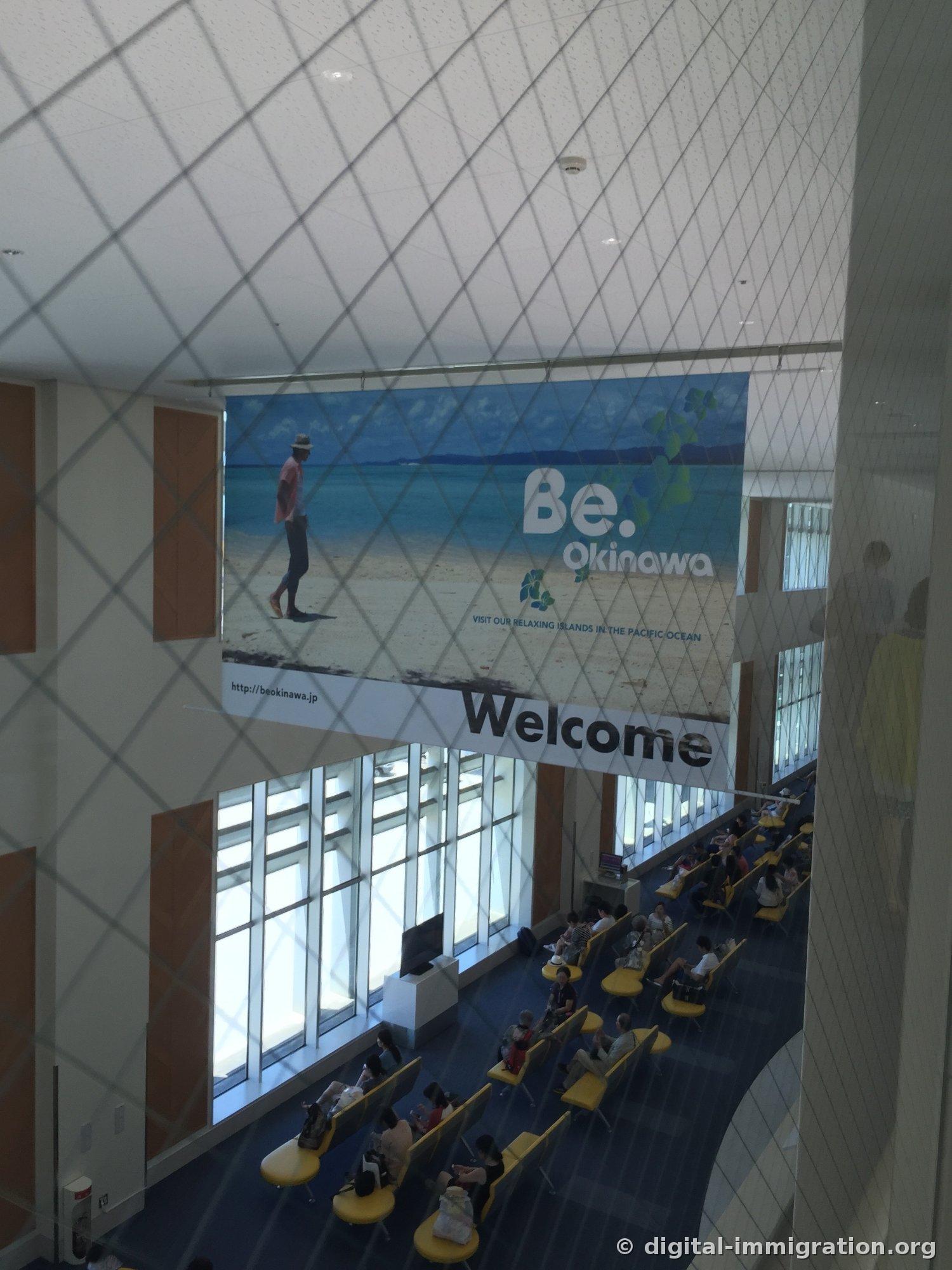Grande bannière 'Be. Okinawa Welcome' suspendue au plafond à l'intérieur du terminal de l'aéroport de Naha. Au-dessous, on voit des passagers assis sur des sièges jaunes en attente, et la lumière du soleil entre par les grandes vitres.