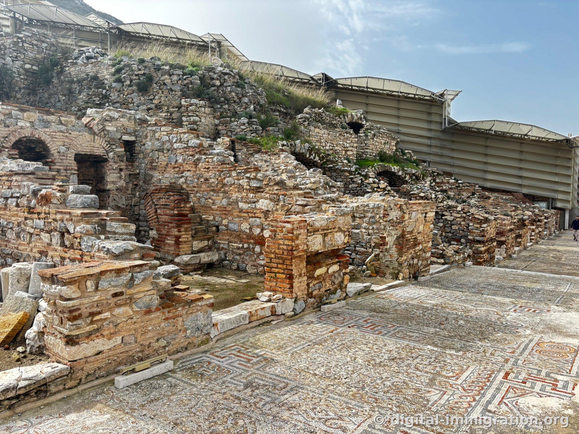 Bathhouse wall made of mixed red brick and stone next to geometric mosaic paving, with a white protective roof visible above on the right
