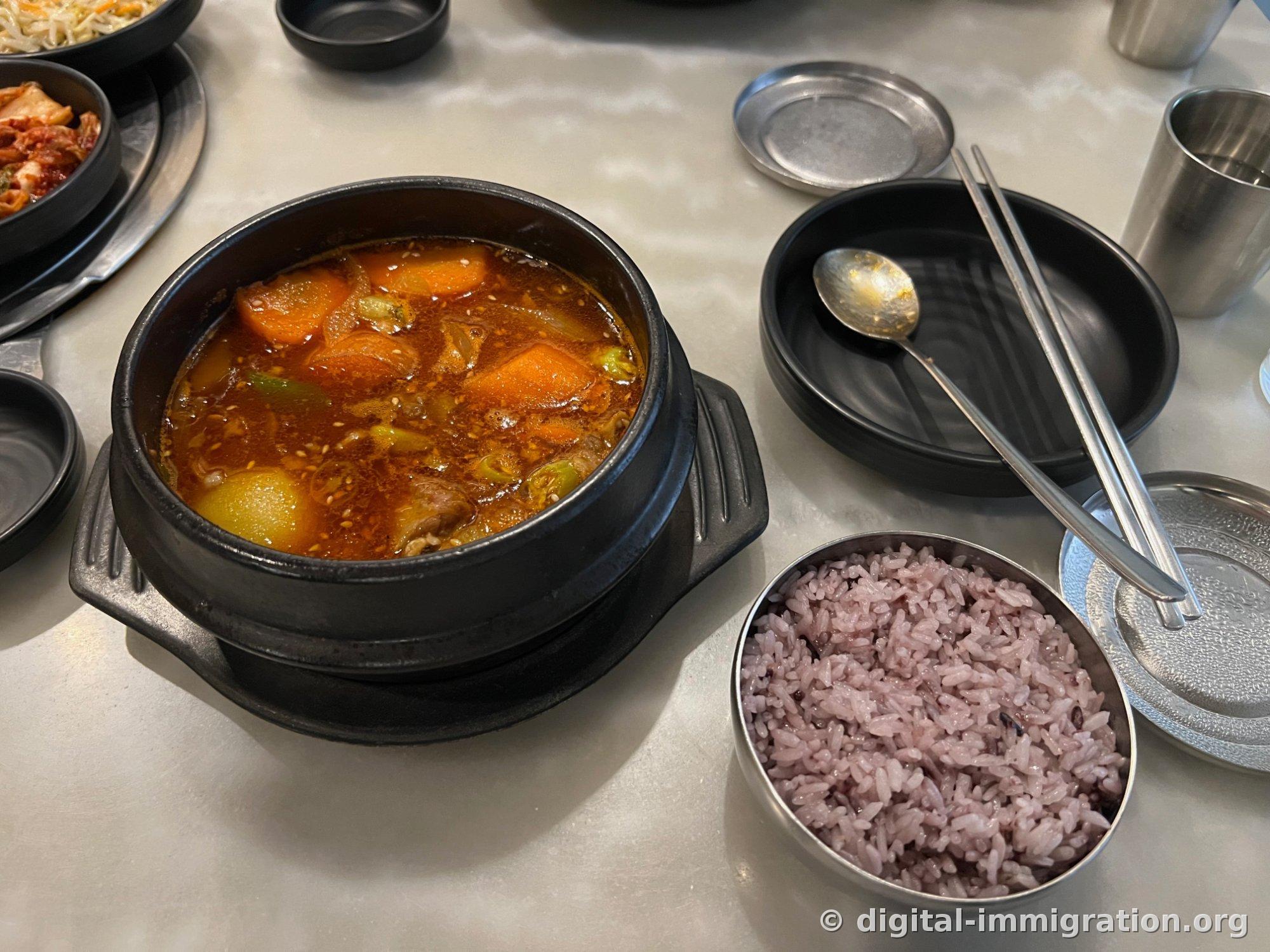 Yukgaejang rouge servi dans un ttukbaegi noir, avec de gros morceaux de carotte et de pomme de terre, du bœuf effiloché en fibres épaisses et du poireau. À côté, un bol en inox de riz noir et des couverts.