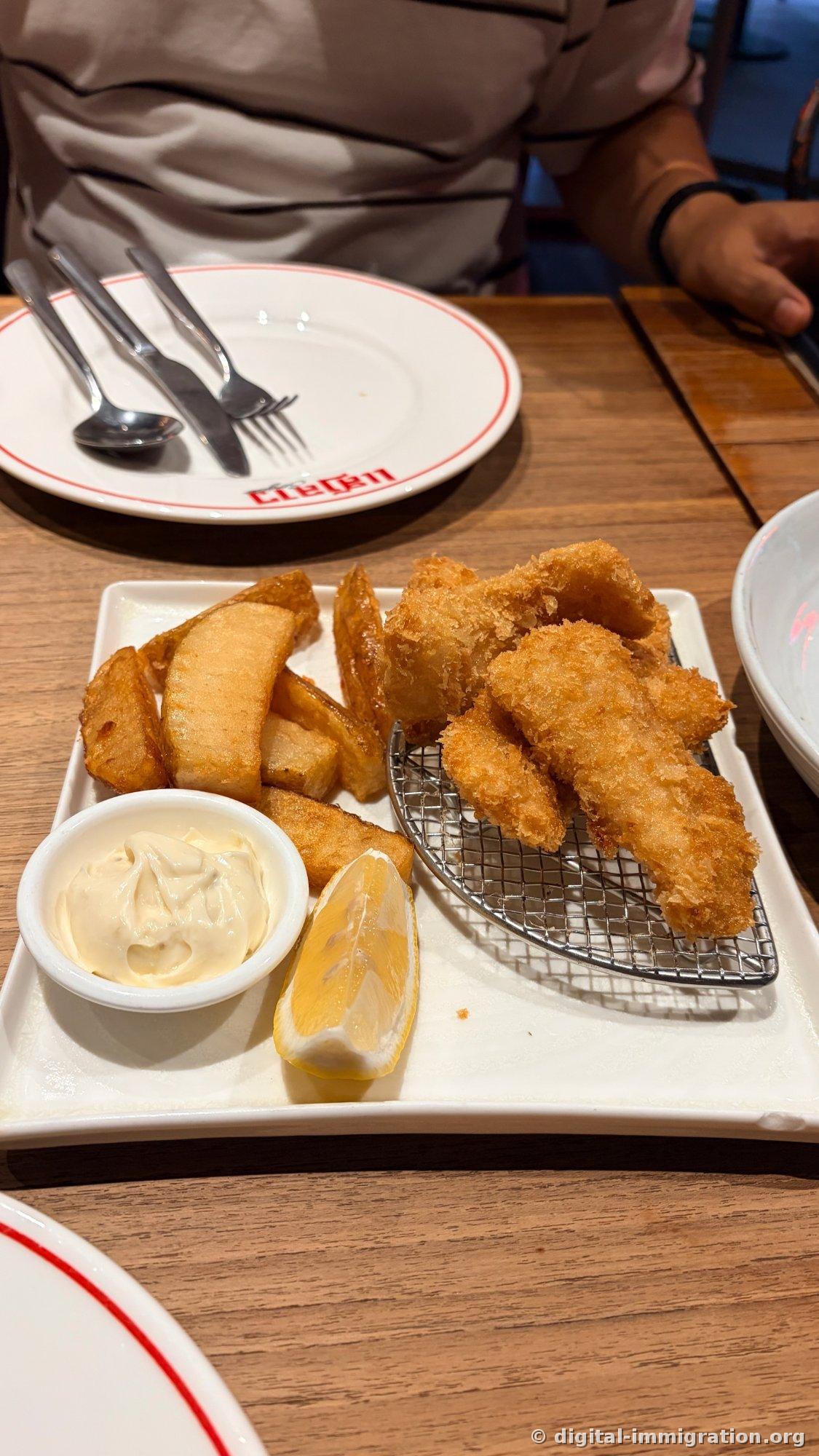 Une assiette blanche carrée avec du poisson frit à la panure et de gros coins de pommes de terre, une sauce tartare et un coin de citron, une assiette ronde avec un logo en laotien derrière