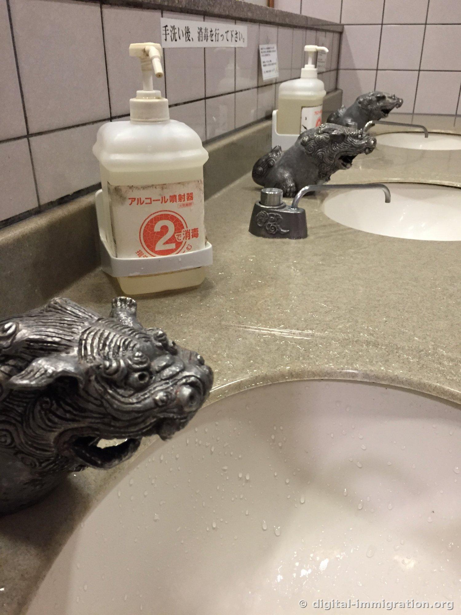 Sink faucet decorated like a shisa with carved mane and teeth, alcohol sanitizer beside it, Japanese instructions on the tiled wall