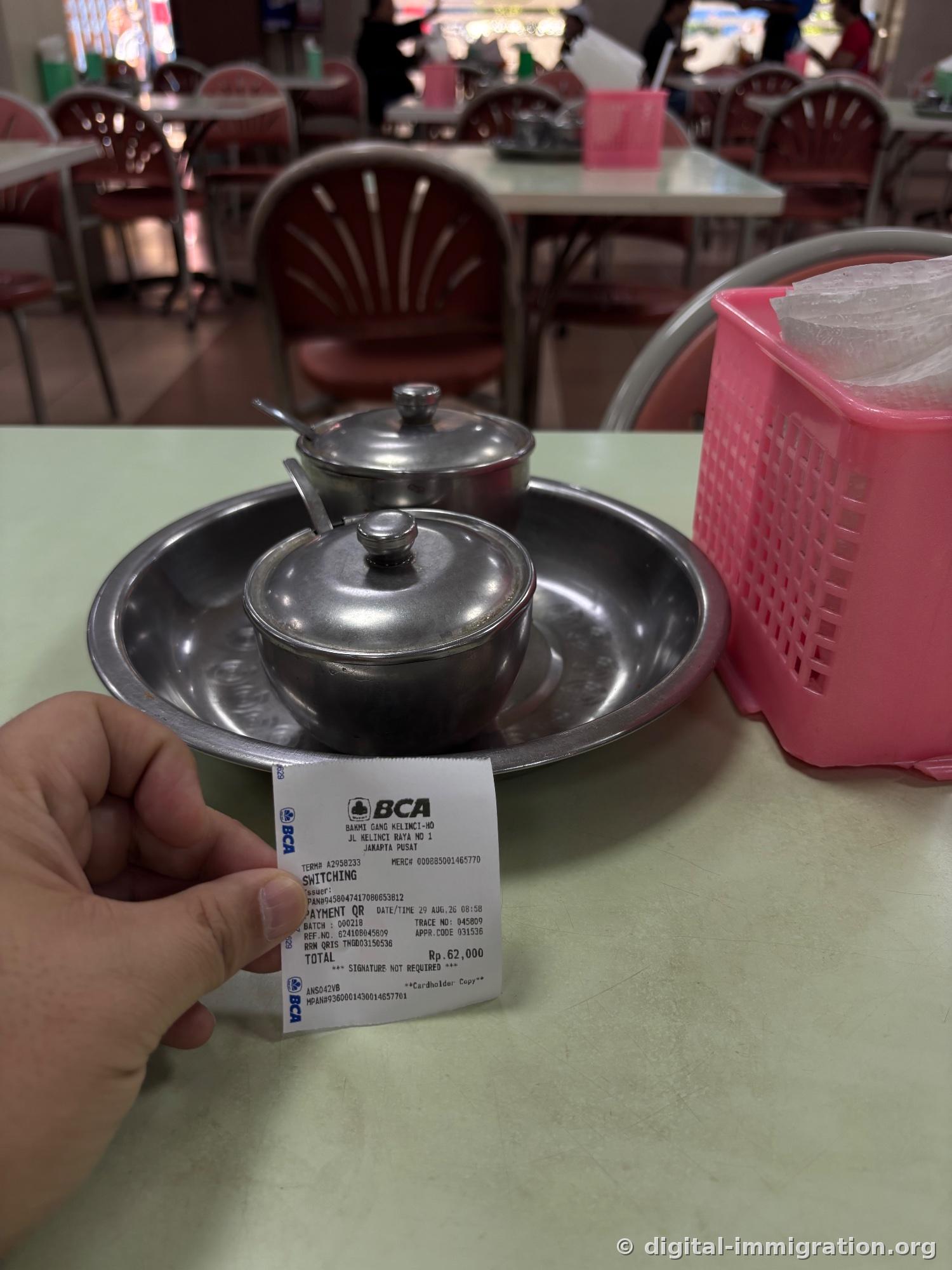 Reçu de carte BCA tenu à la main affichant 'BAKMI GANG KELINCI-HO, JL KELINCI RAYA NO 1, JAKARTA PUSAT', 29 AUG 26 08:58, TOTAL Rp.62,000, avec pots de sauce et distributeur de serviettes rose en arrière-plan