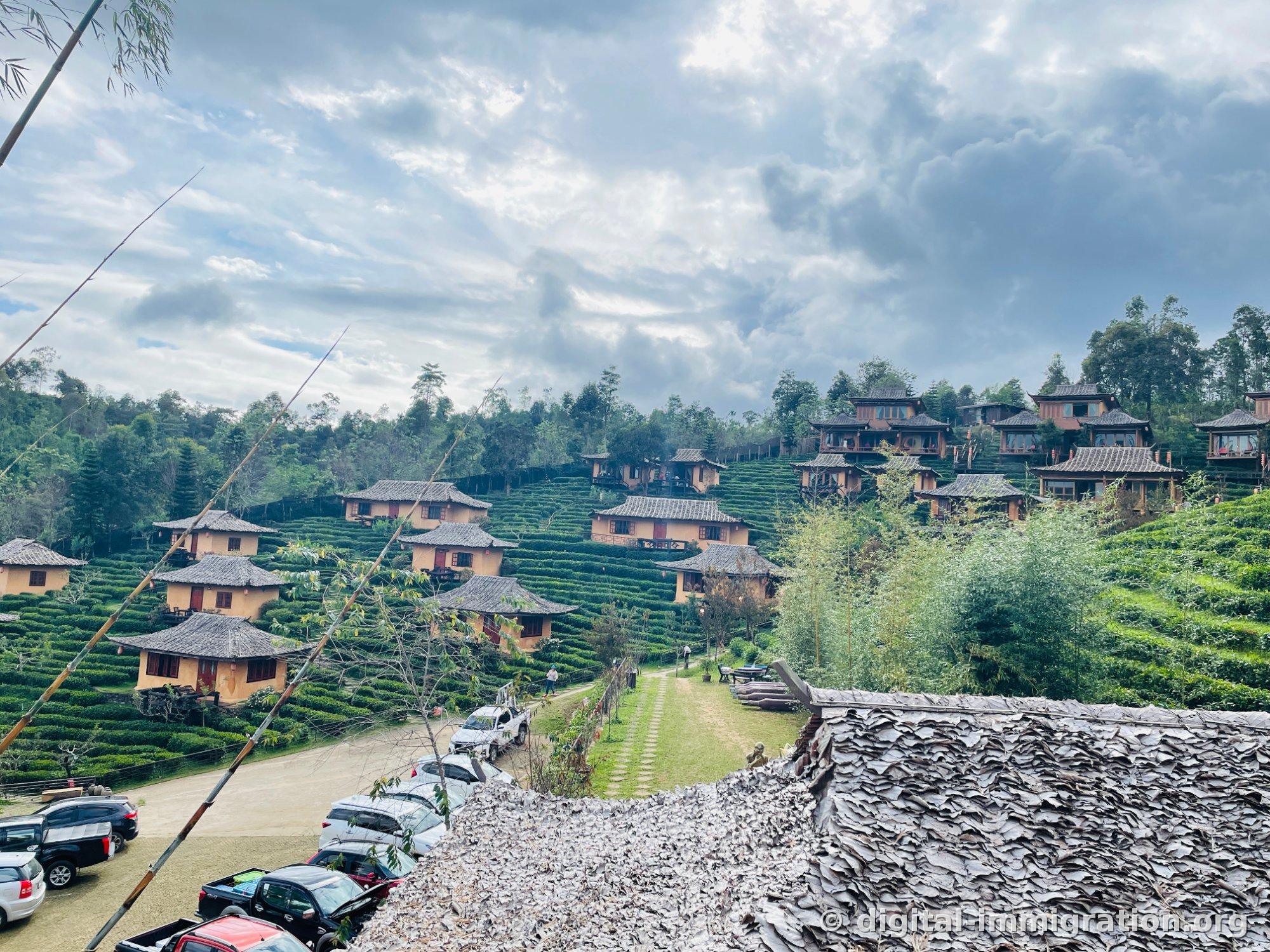 Petites maisons d'hôtes en terre couleur ocre sur la colline de Ban Rak Thai avec des plantations de thé en terrasses. Des séchoirs de feuilles gris au premier plan et des véhicules garés