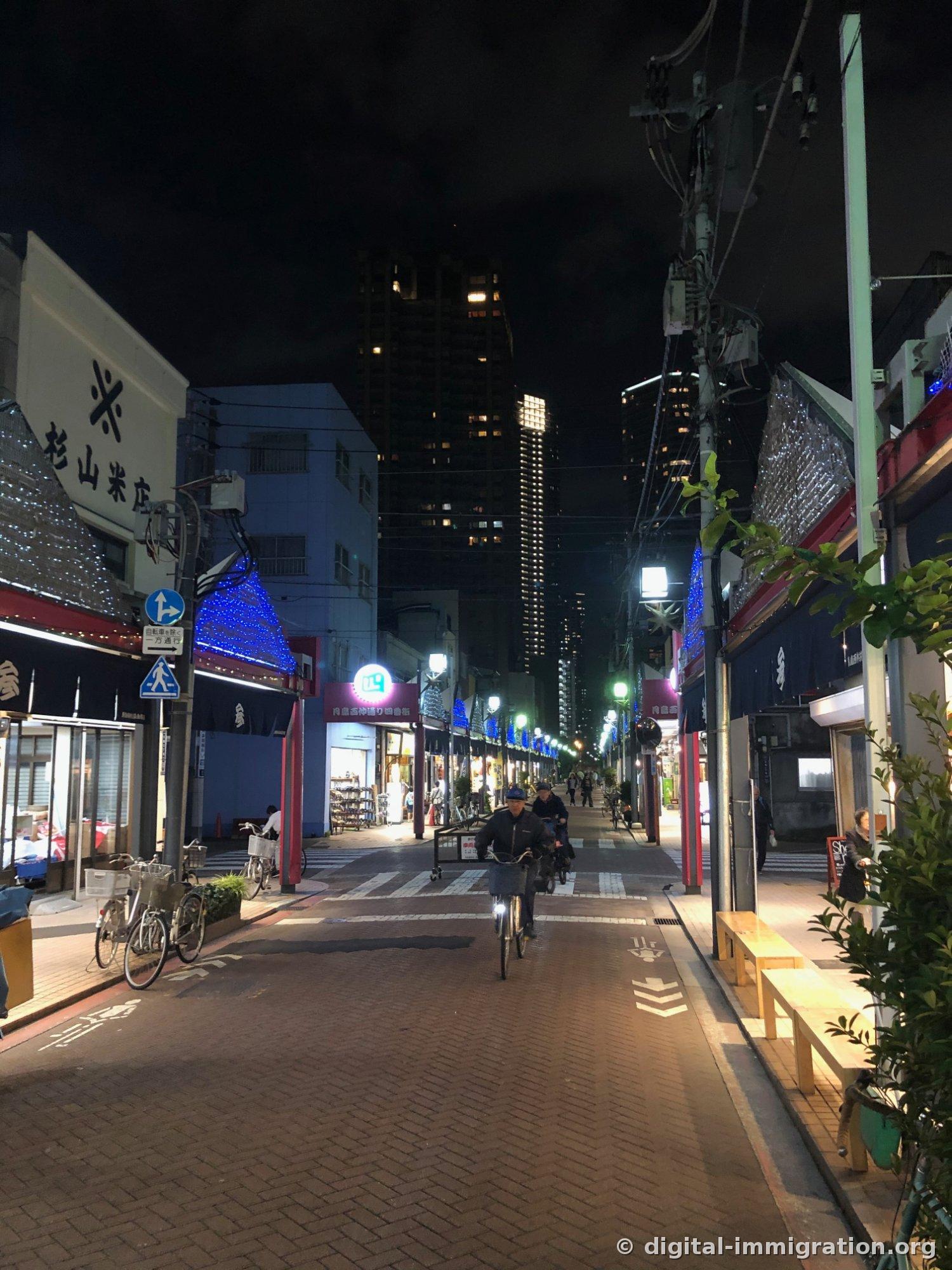 '杉山米店' 간판과 츠키시마 니시나카도리 상점가 표지, 자전거를 타고 다가오는 주민, 뒤편에 고층 타워맨션