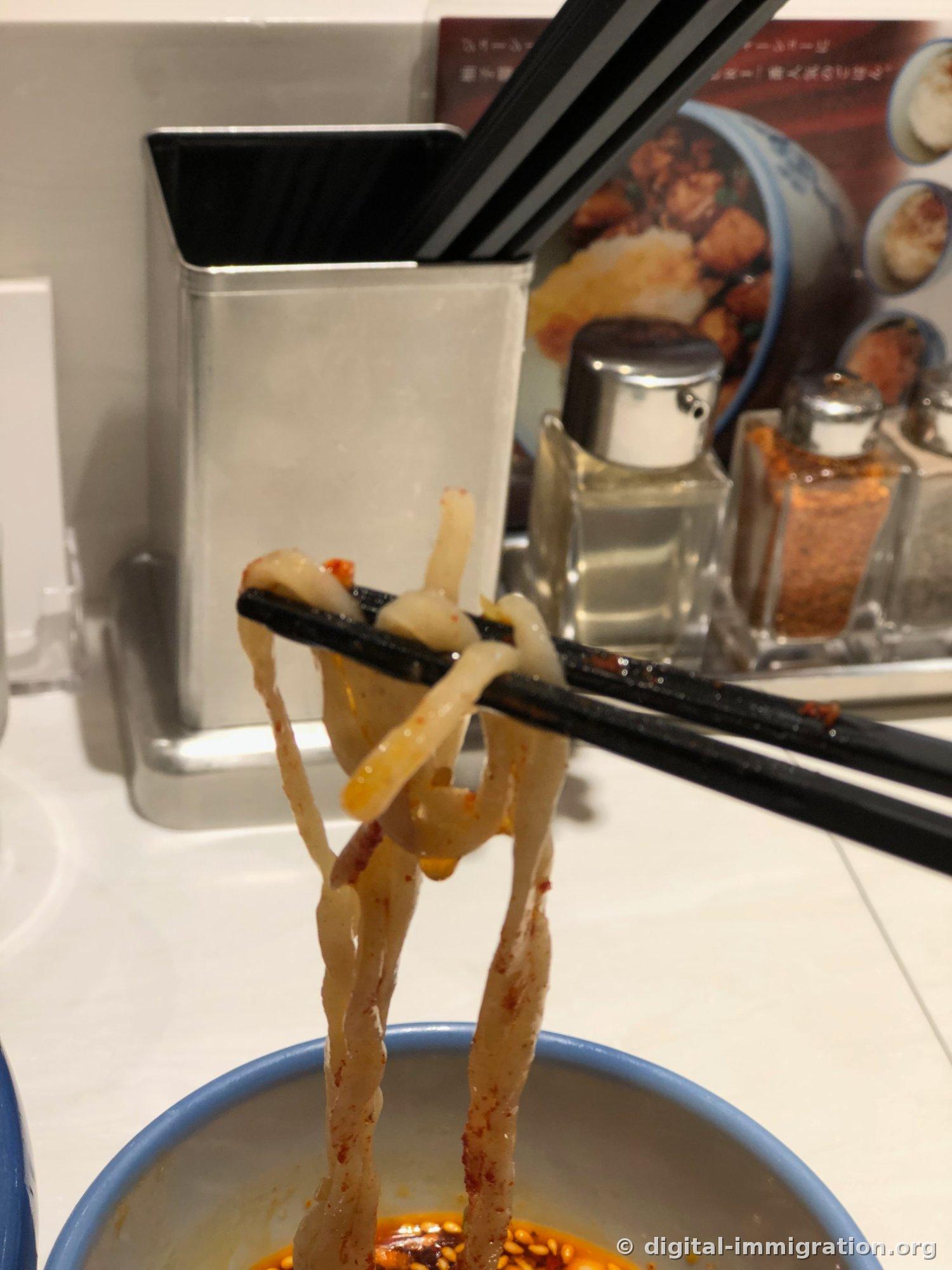 Thickly coated noodles lifted high with black chopsticks, seasoning bottles for garlic oil and chili powder visible at the back table