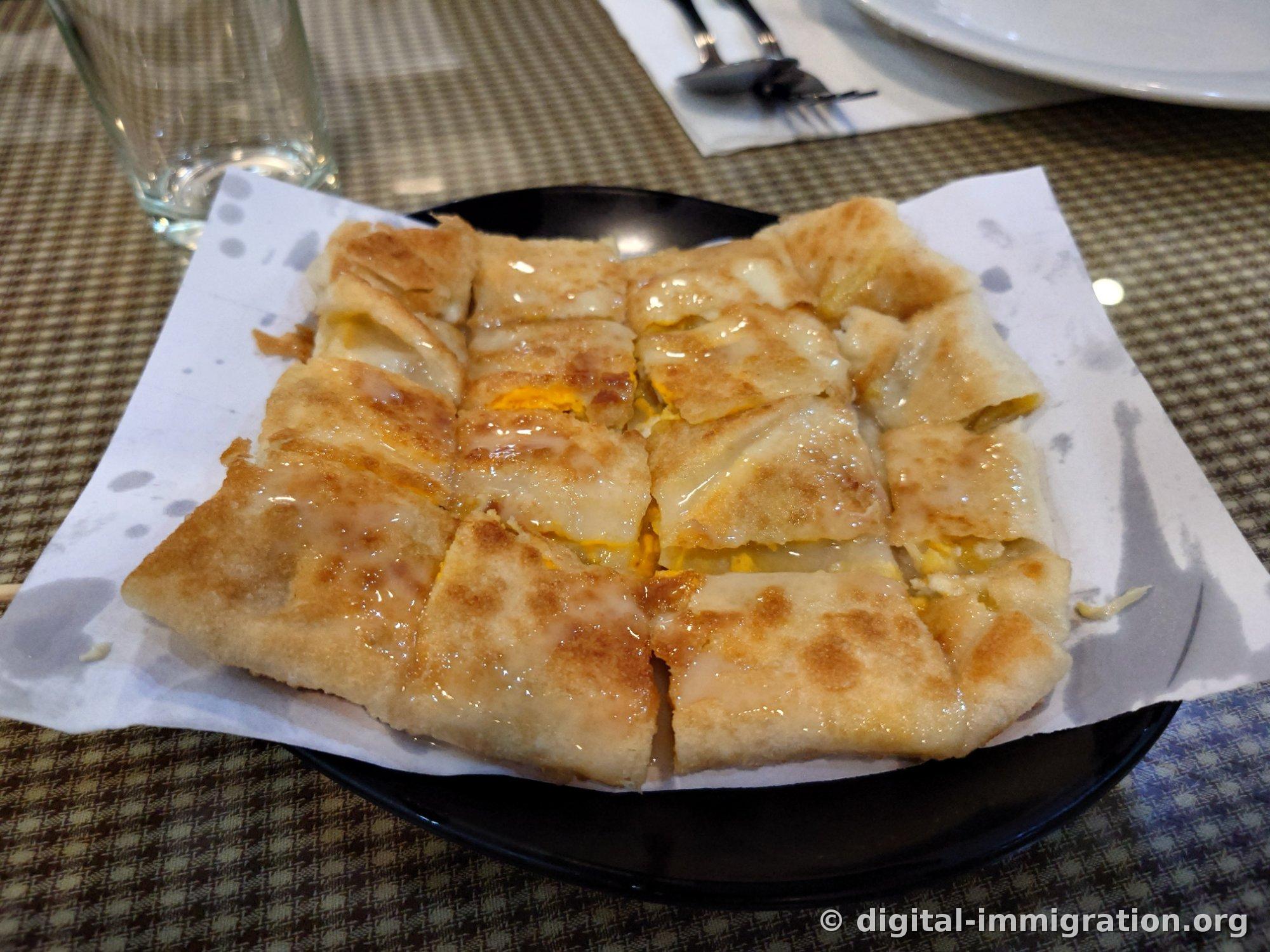 Banana roti cut into squares on paper-lined black plate, with condensed milk gleaming on the surface