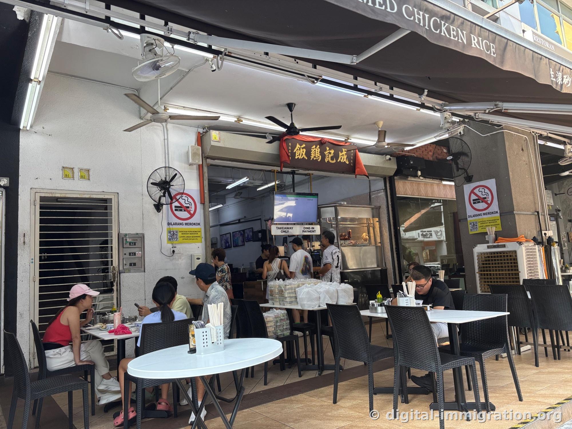 Customers dining at outdoor tables under the STEAMED CHICKEN RICE canopy, with takeaway bags piled at the counter inside