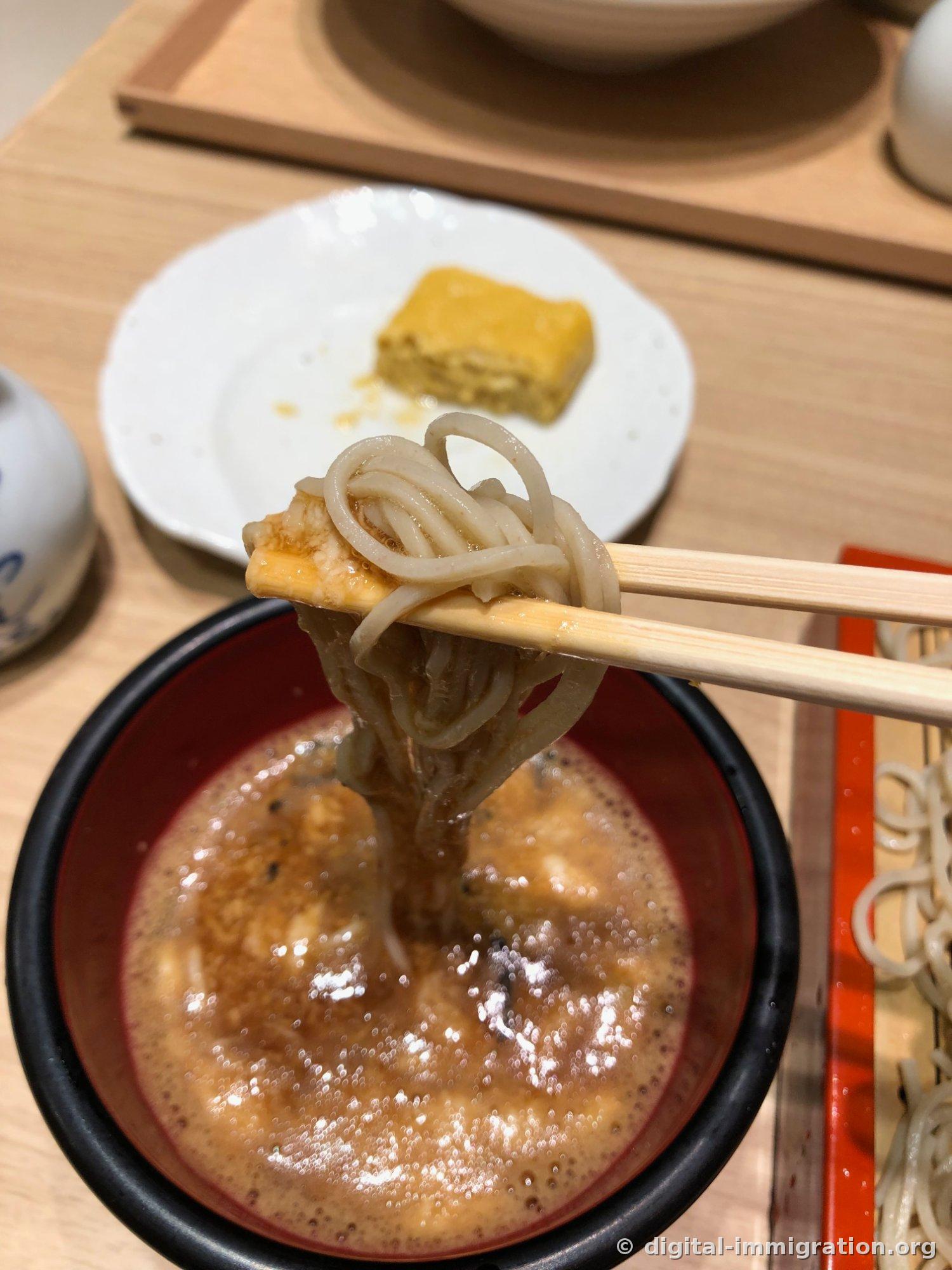 Lifting soba noodles with chopsticks from the foamy brown sauce of mixed tororo, egg yolk, and tsuyu