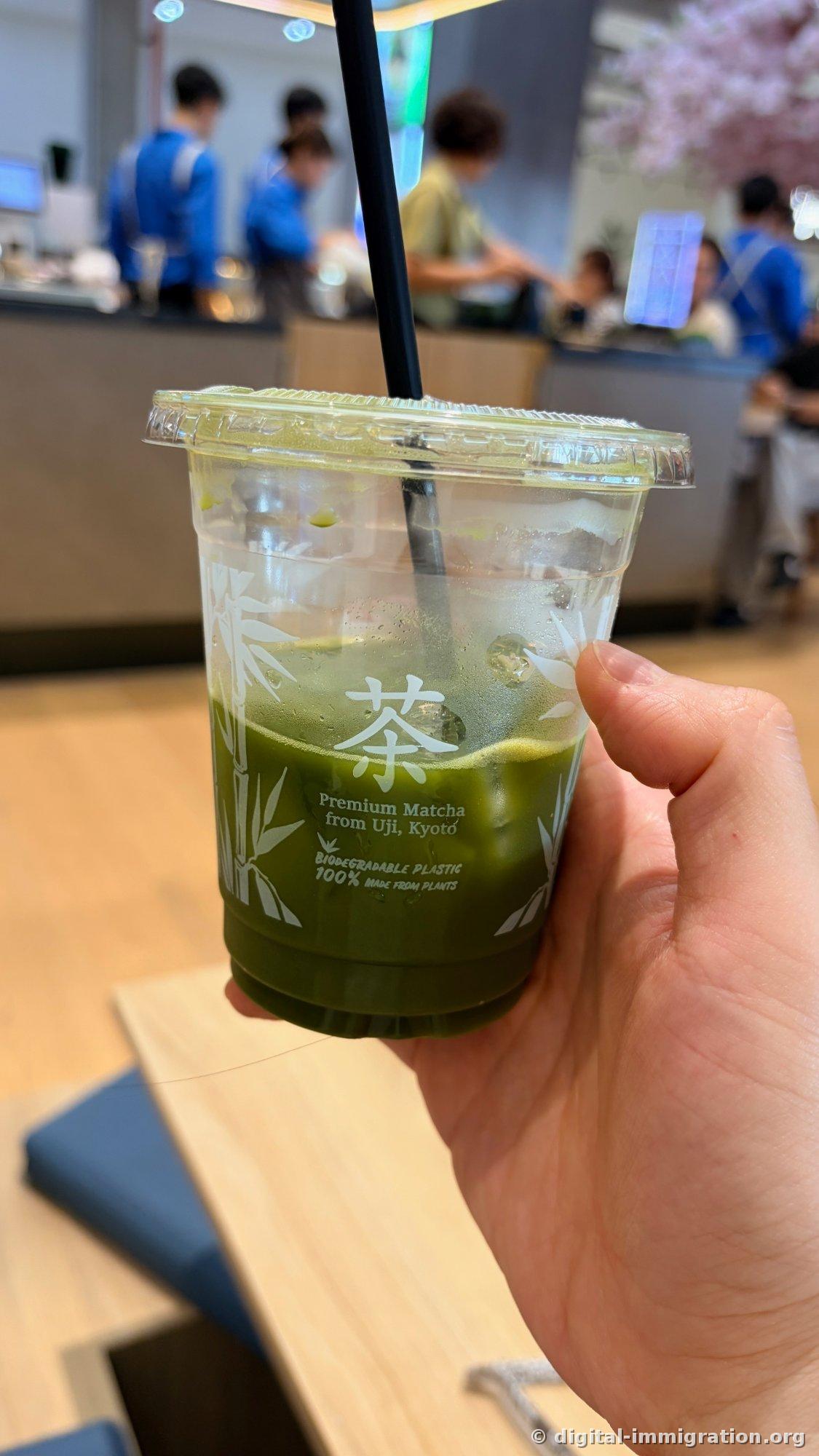 Iced matcha in a clear plastic cup held in hand, with '茶' character and 'Premium Matcha from Uji, Kyoto' text on the cup, staff members in blue uniforms and artificial cherry blossoms blurred in the background