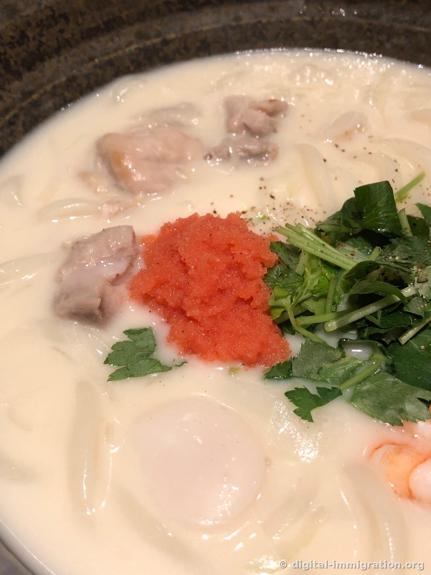 Close-up photo of cream udon, with red cod roe grains, green mitsuba, chicken chunks, and scallops visible on the creamy white broth