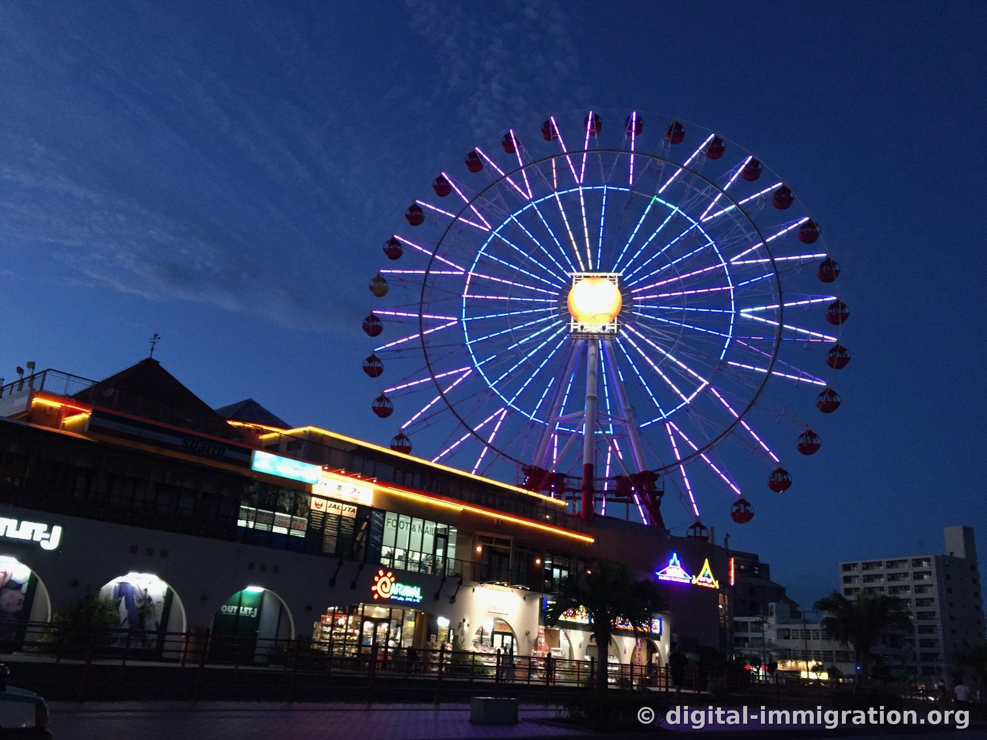 American Village's iconic observation wheel glowing with purple and blue LEDs against the deep blue twilight sky, with the OUTLET-J and Carnival Park signs lit up on the shopping mall below
