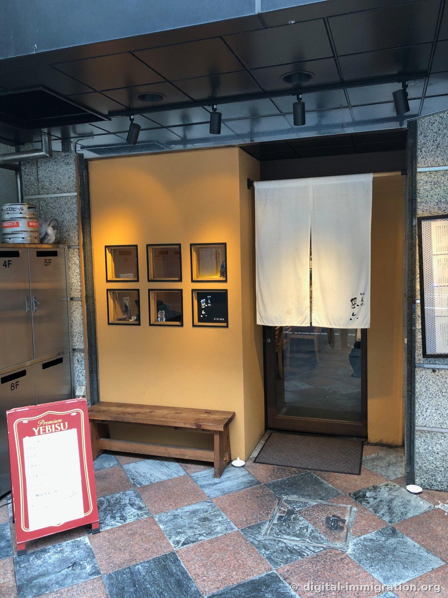 ビル1階に白い暖簾が掛かった甚六の入口、黄色い壁に黒い額の銘板と木製ベンチ、隣には赤いエビスビールの看板が立っている