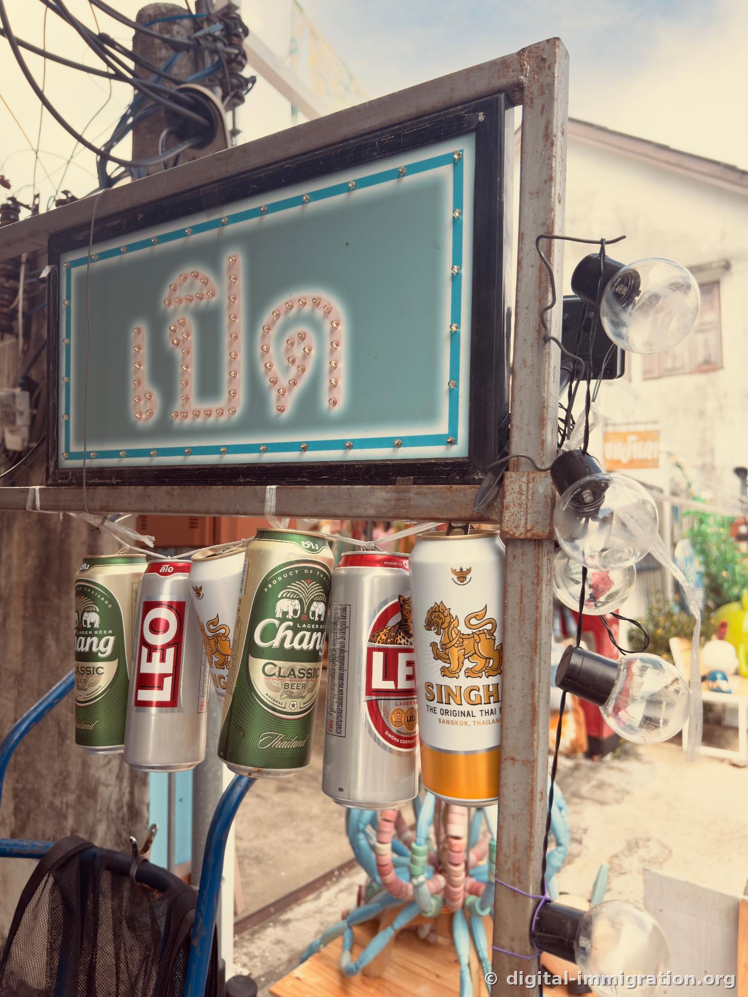 Thai language sign with light bulbs and hanging empty beer cans of Chang, Leo, Singha brands below, tangled power lines on utility pole in background