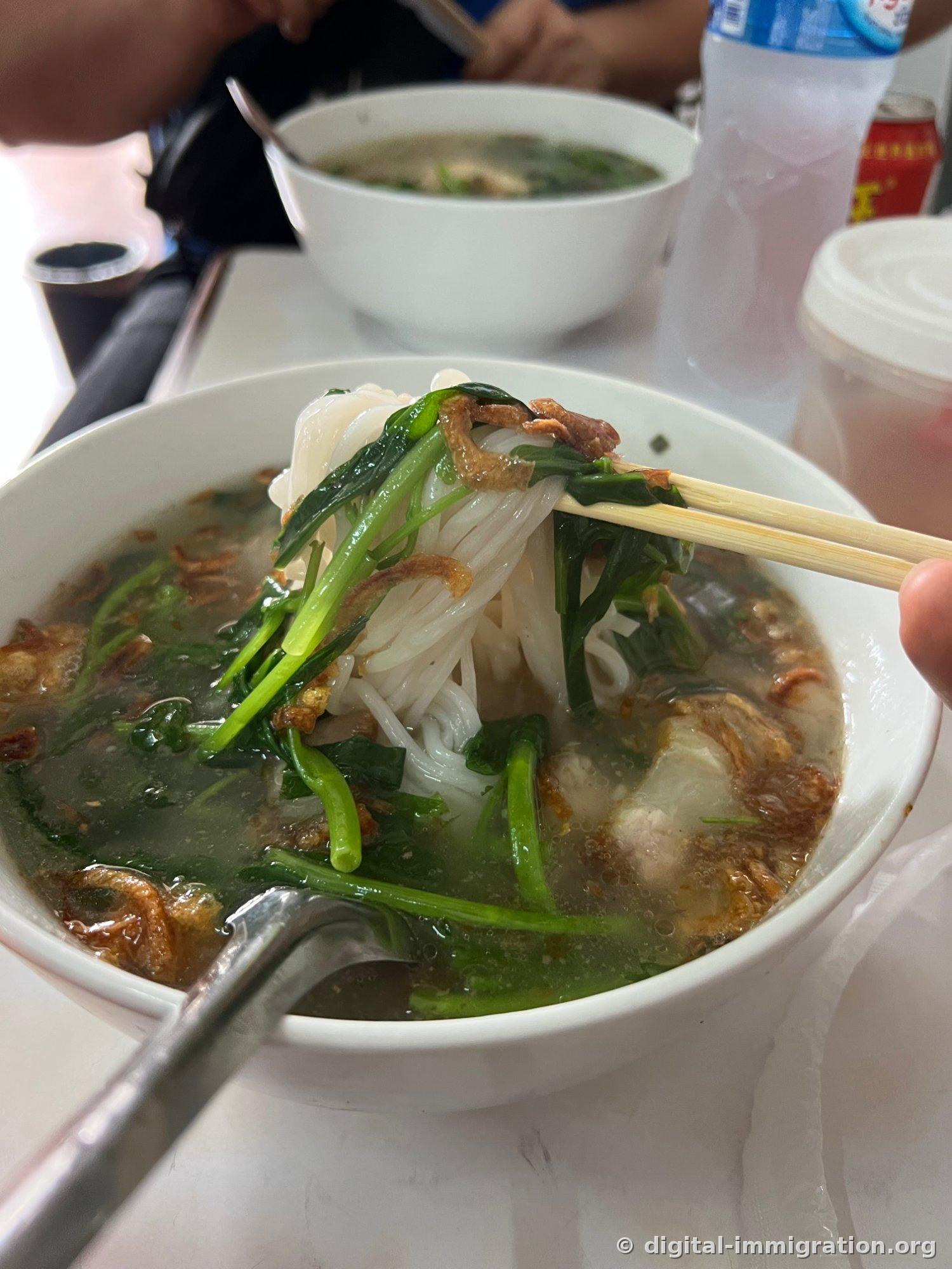 White rice noodles lifted from thick broth with wooden chopsticks, with green vegetables and fried garlic clinging to the noodles, and another bowl of noodles and a water bottle on a table in the background