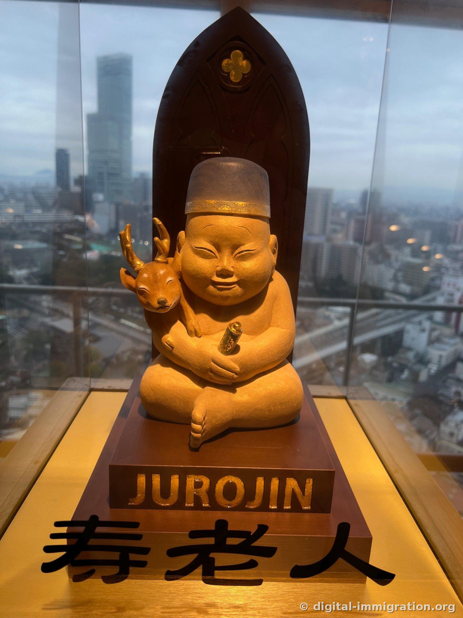 A wooden statue of Jurōjin sitting and holding a deer, with Abeno Harukas visible outside the window