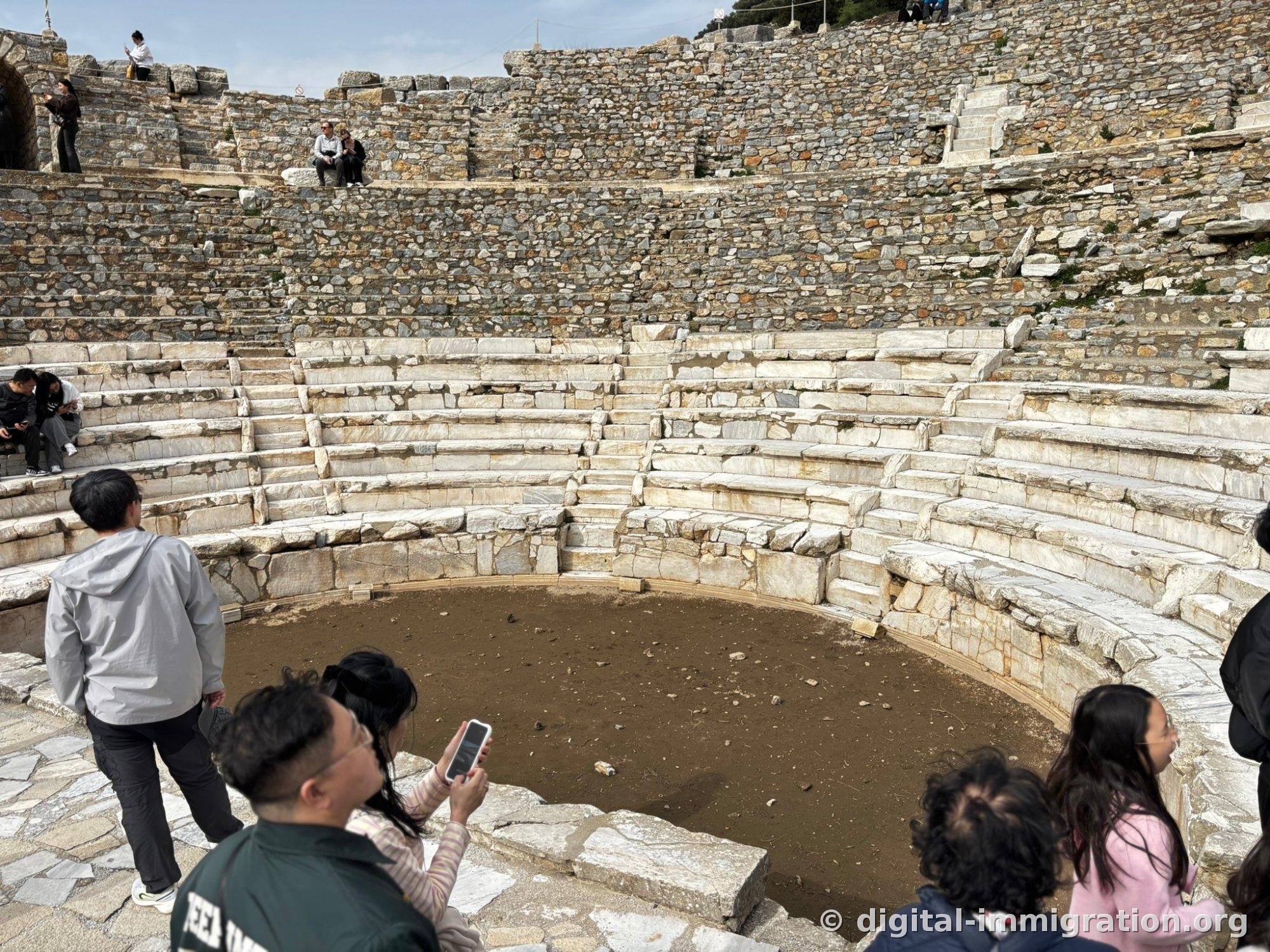 Tourists sitting in the semicircular audience seating of the Ephesus Odeum with the orchestra floor below covered in soil