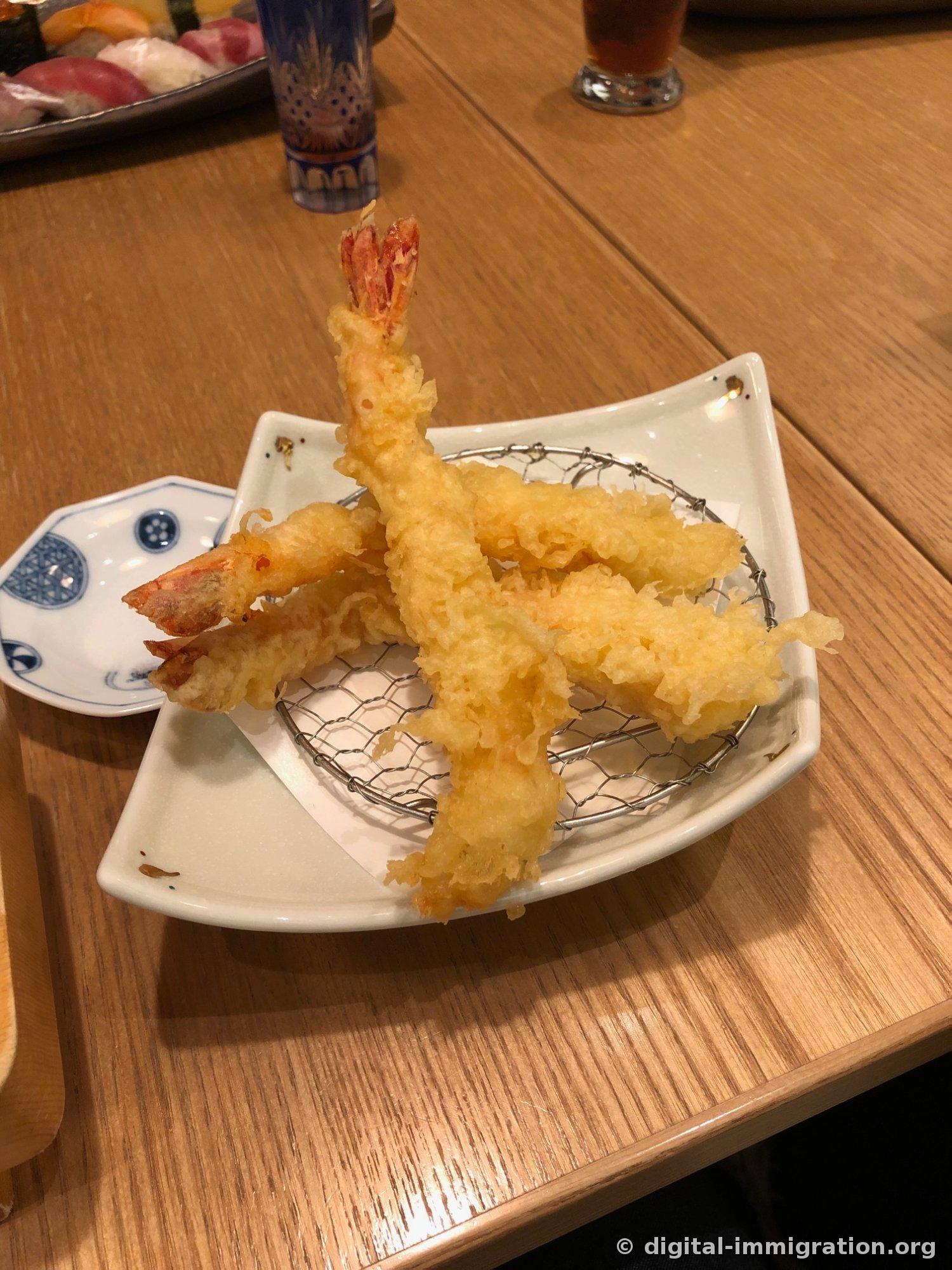 Trois crevettes tempura dressées queue vers le haut sur un filet métallique dans une assiette blanche carrée. Derrière, on voit le plateau de sushis et un verre de thé glacé