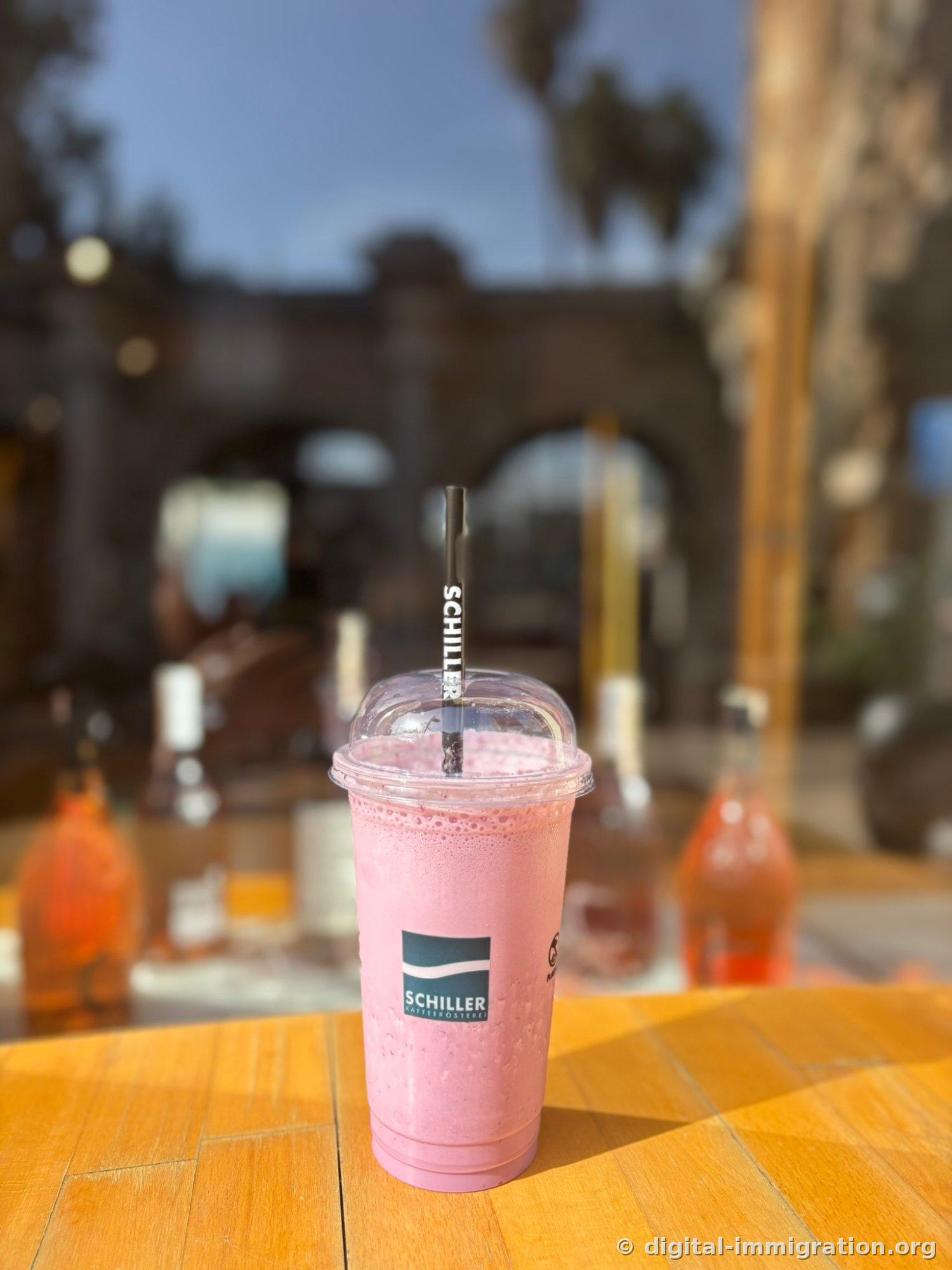 Un verre transparent avec le logo SCHILLER posé sur une table en bois, contenant un smoothie aux fraises rose. Une paille noire avec le texte SCHILLER, en arrière-plan un bâtiment en pierre voûtée flou