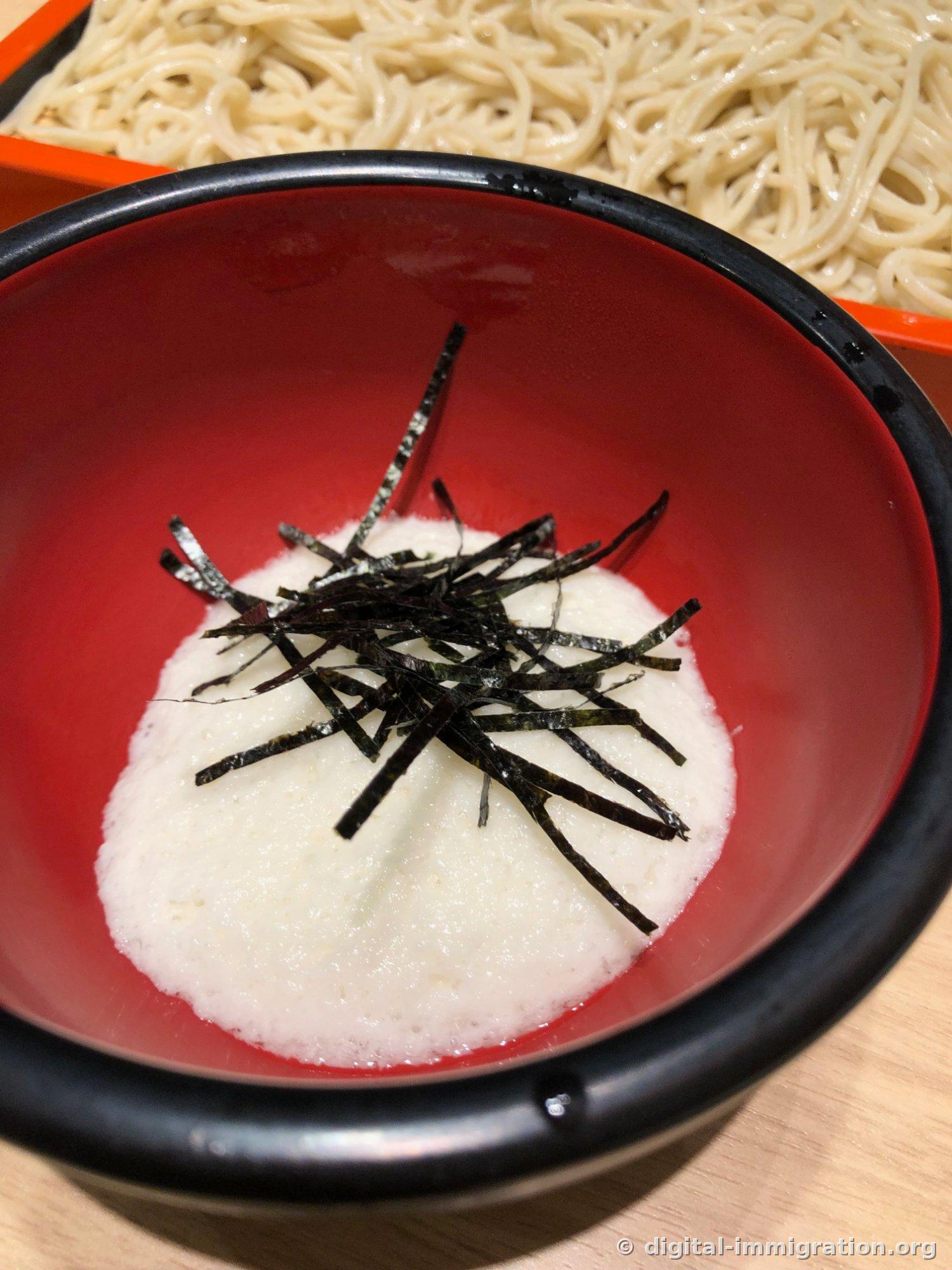 A dark reddish bowl of thick, mounded tororo with shredded nori on top, with soba visible in the background