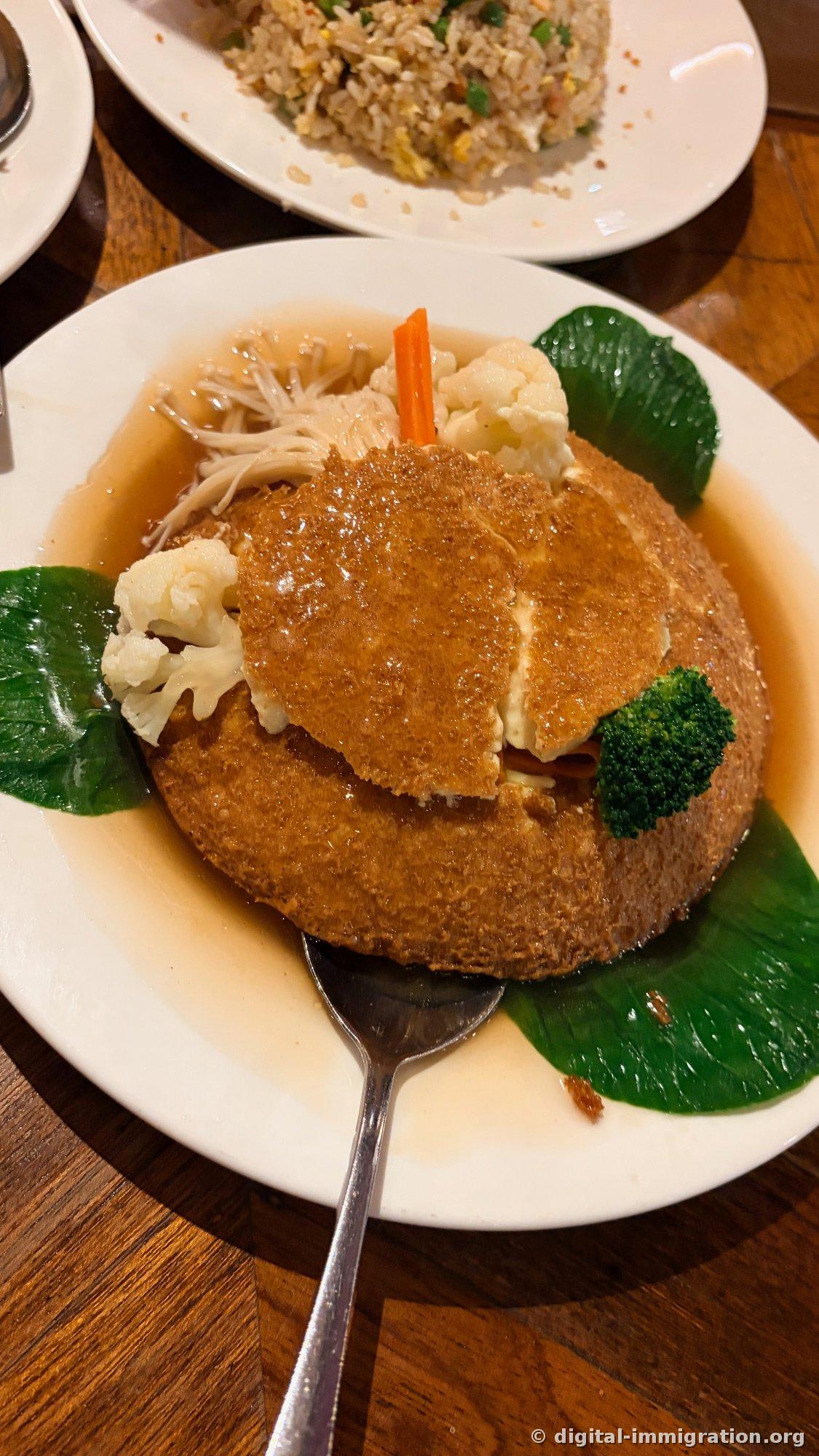 A thick fried tofu disc served in light brown soy broth, surrounded by enoki mushrooms, cauliflower, broccoli, bok choy, and carrot