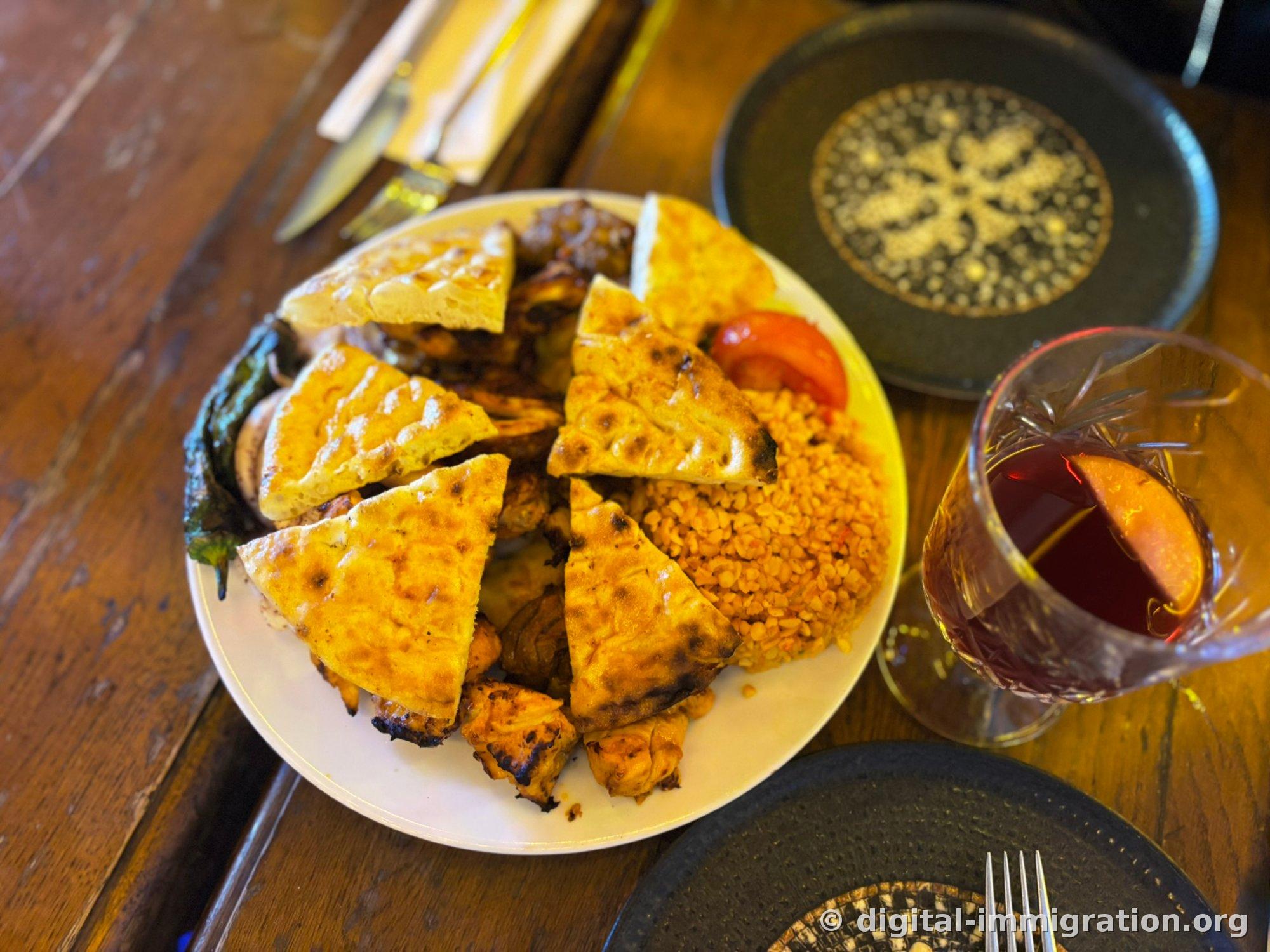흰 접시에 부채꼴로 자른 구운 라바쉬와 닭 시시케밥, 붉은 불구르 필라프, 구운 토마토와 청고추가 담겨 있고 옆에 뱅쇼 잔이 놓여 있다