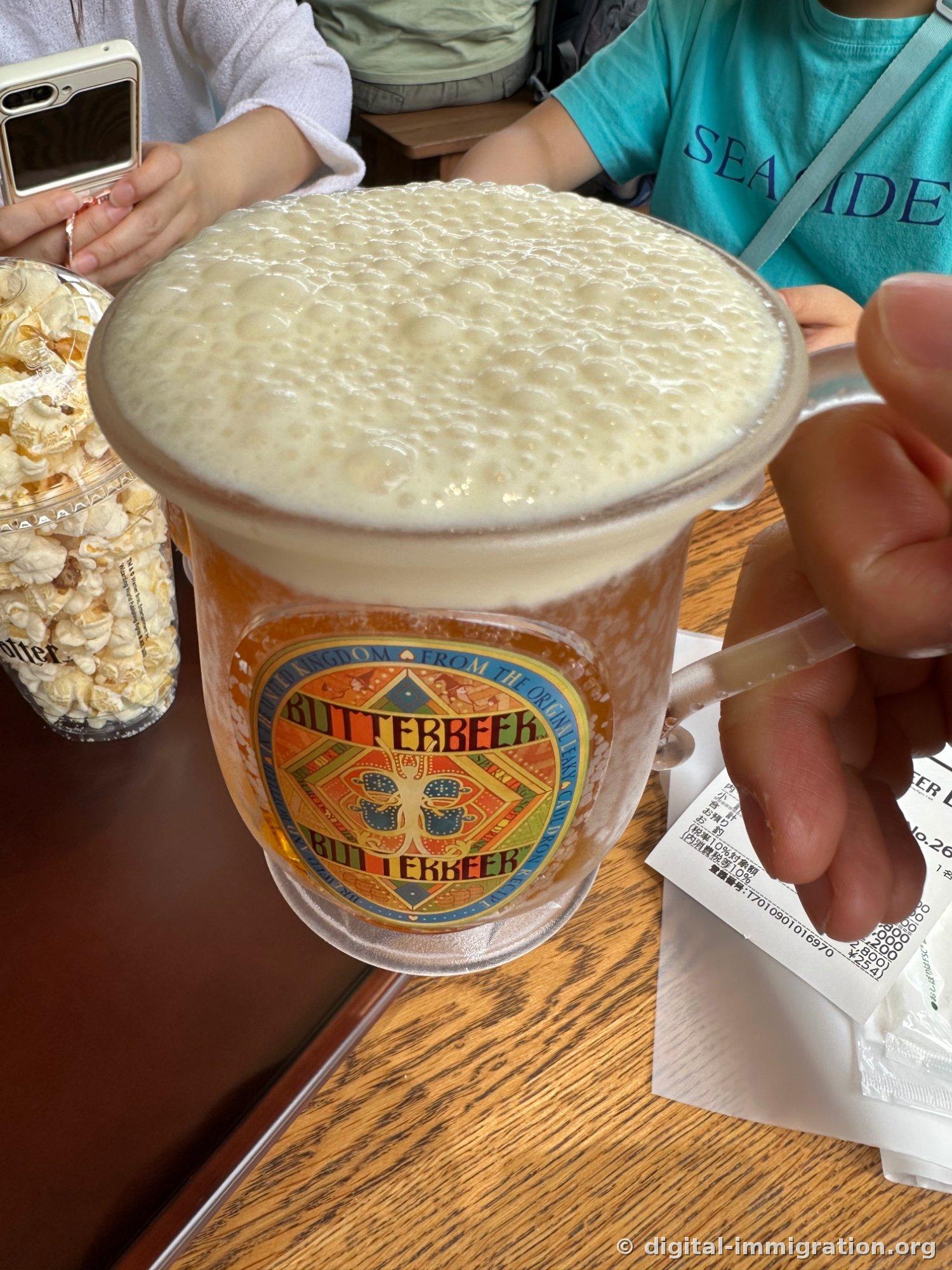A glass with a BUTTERBEER label and handle topped with thick foam, next to a popcorn cup