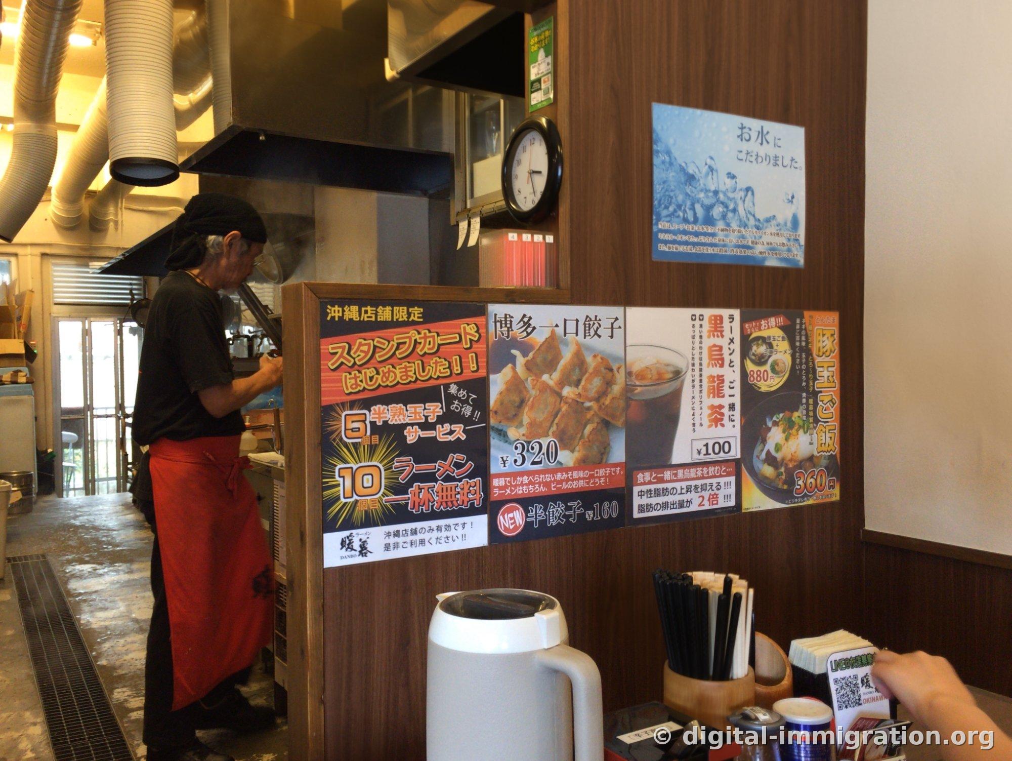 L'intérieur du restaurant Tanpo. Un employé en foulard noir et tablier rouge travaille dans la cuisine, au-dessus du comptoir un avis de carte de fidélité et des affiches pour gyoza 320 ¥, thé oolong noir 100 ¥, riz butakotama 360 ¥