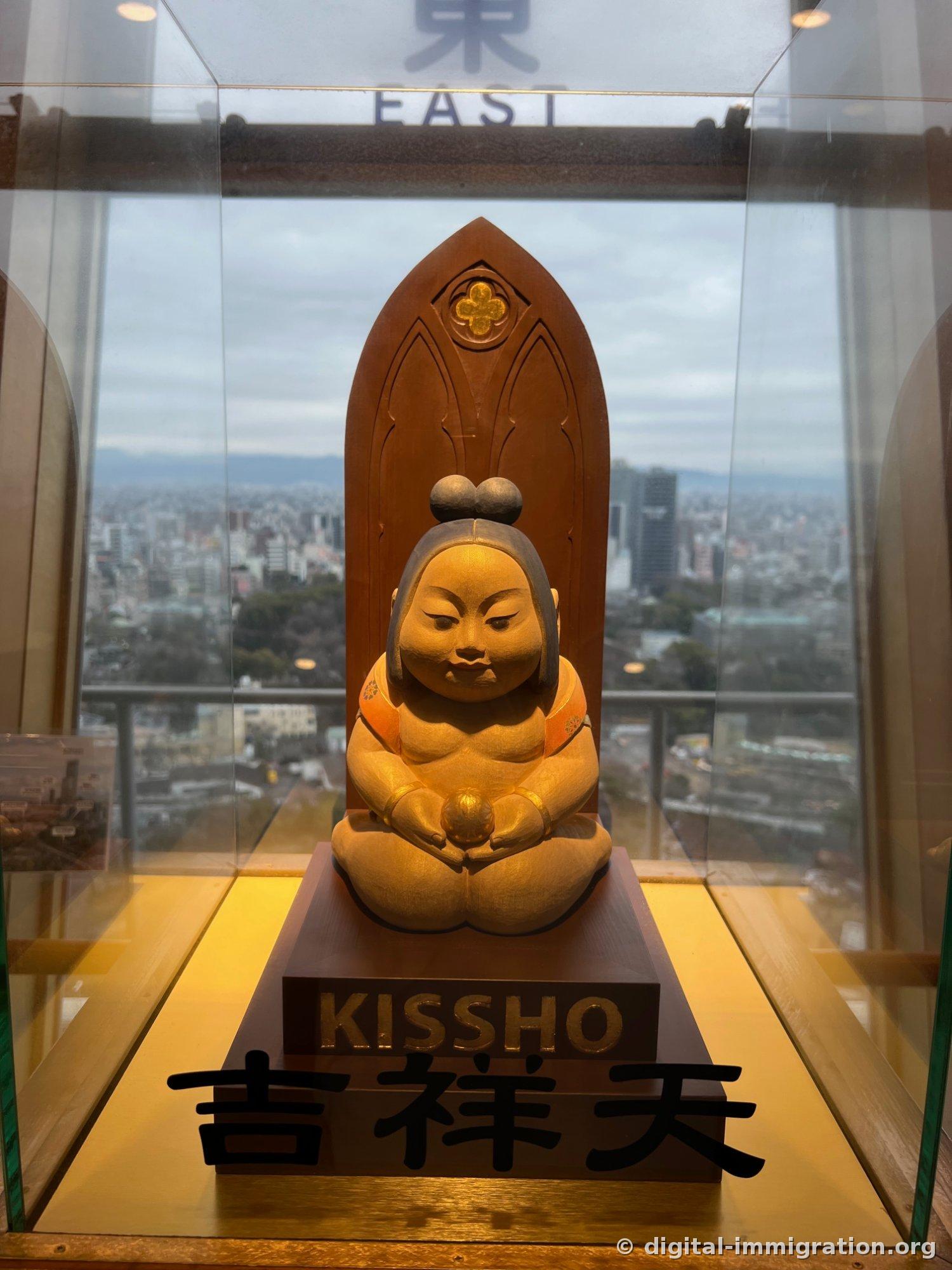 A wooden statue of Kichijoten in a glass case, with the EAST direction indicator and Osaka city in the background
