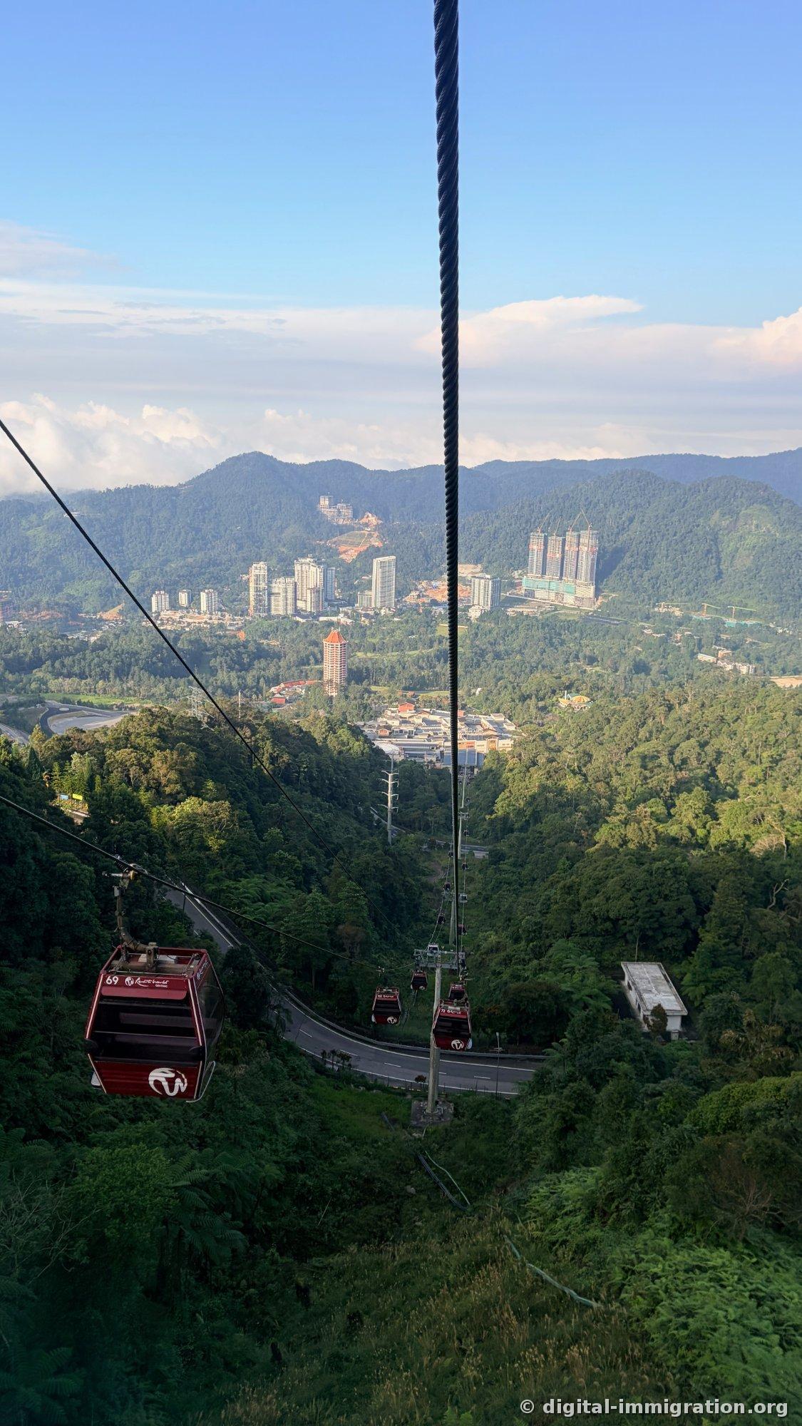 View from the cable car showing cabin 69 passing by, winding road between rainforest, and Genting city high-rises in the distance