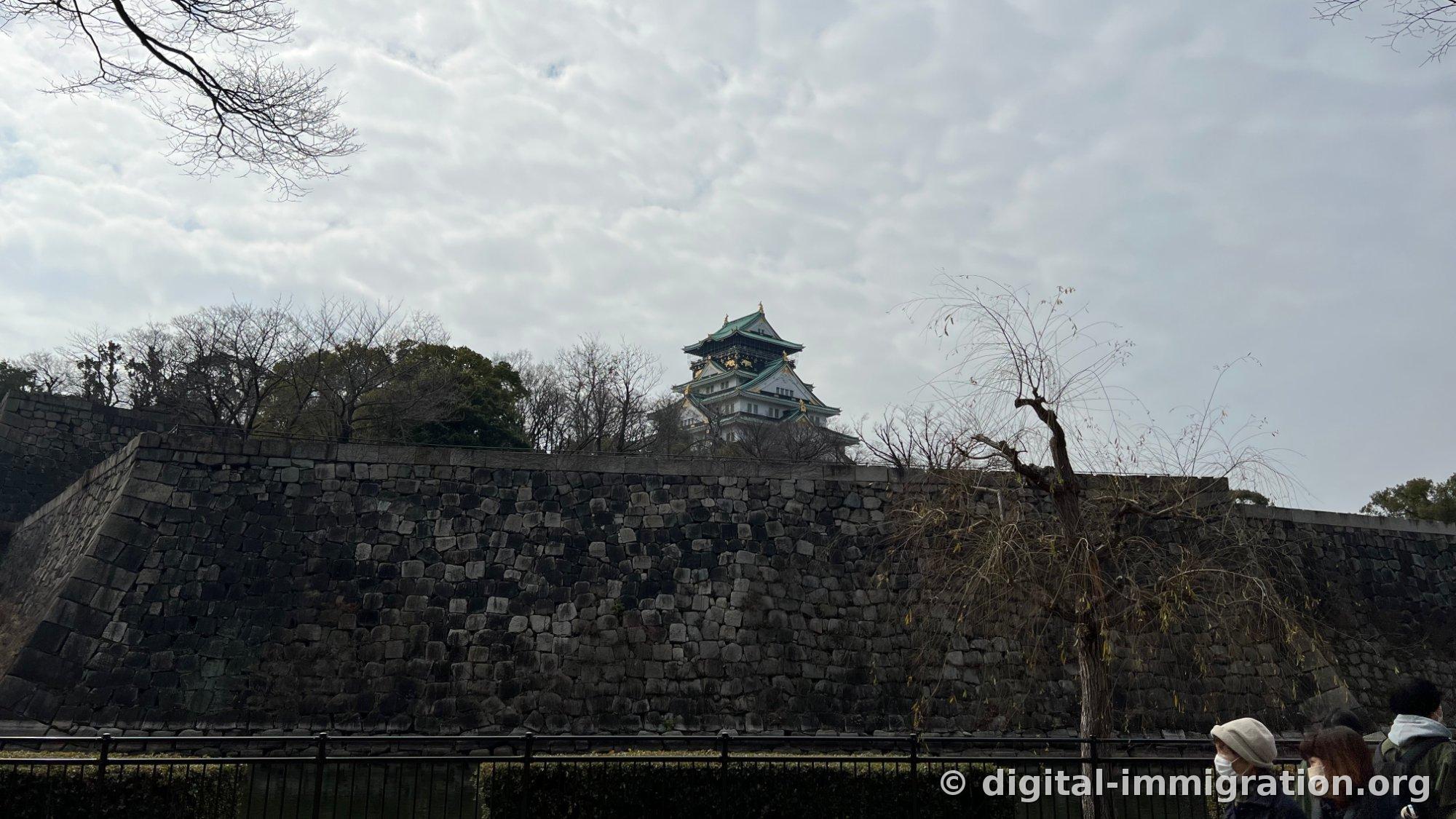 Silhouette de la tour de guet du château d'Osaka se dressant au-dessus du long mur de pierre sous un ciel couvert, avec une rampe en métal noir et des touristes au premier plan