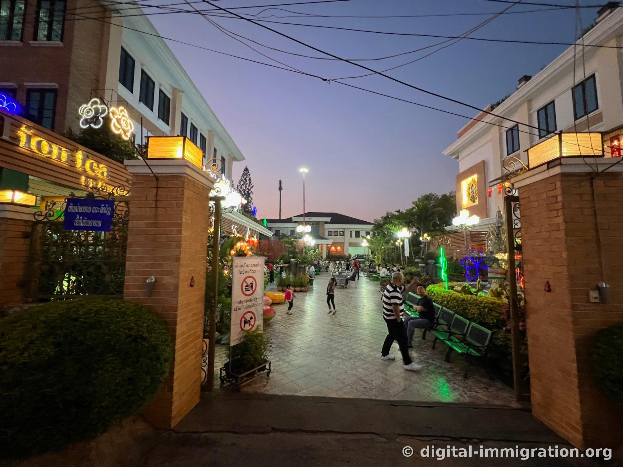 The main entrance of Poysian with flower neon lights illuminated between brick pillars under the darkening sky, with customers seated in the courtyard inside