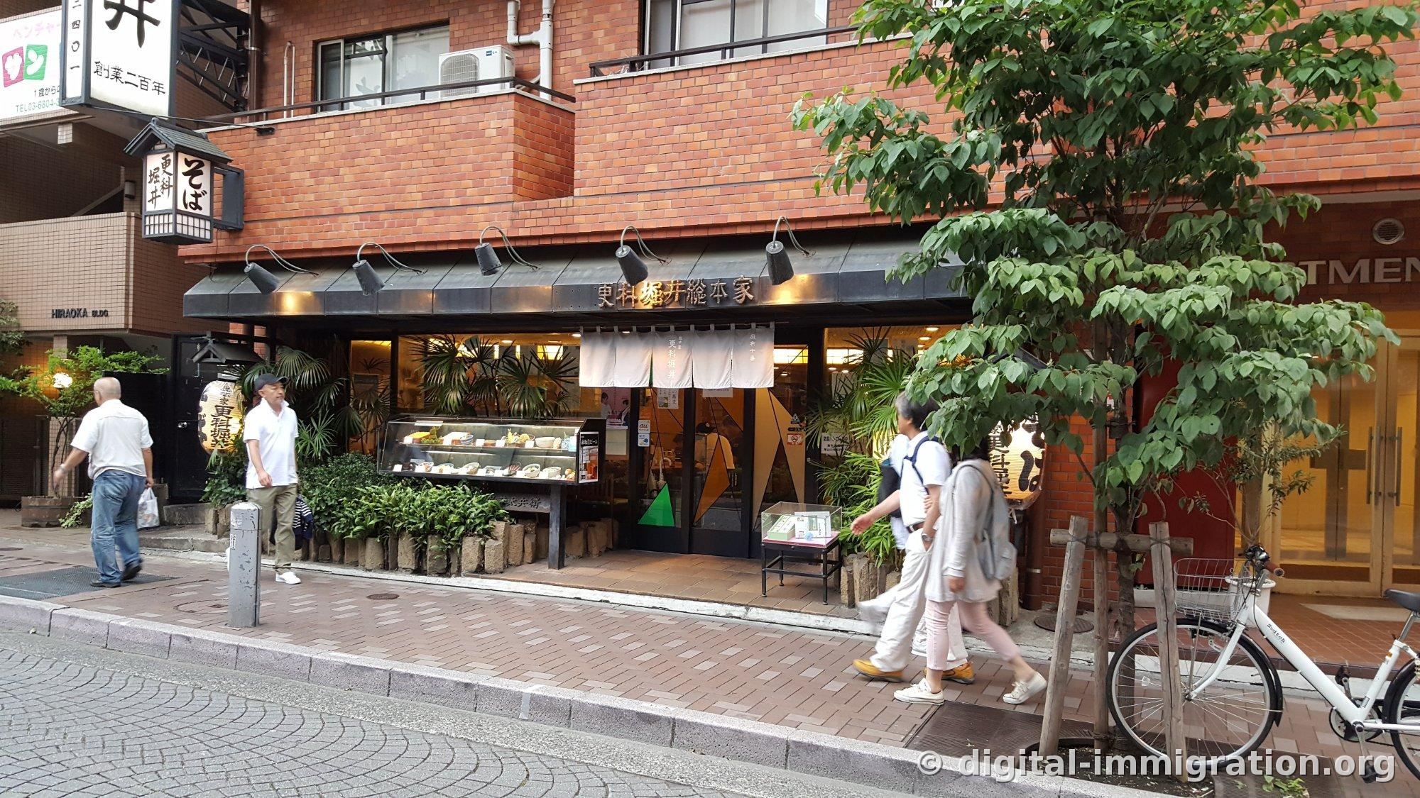 Façade du Sarashina Horii Sohonke au premier étage d'un bâtiment en briques rouges dans une ruelle d'Azabu-Juban, avec la lanterne en bois et le noren blanc, la vitrine avec les modèles de plats, et des piétons passant devant