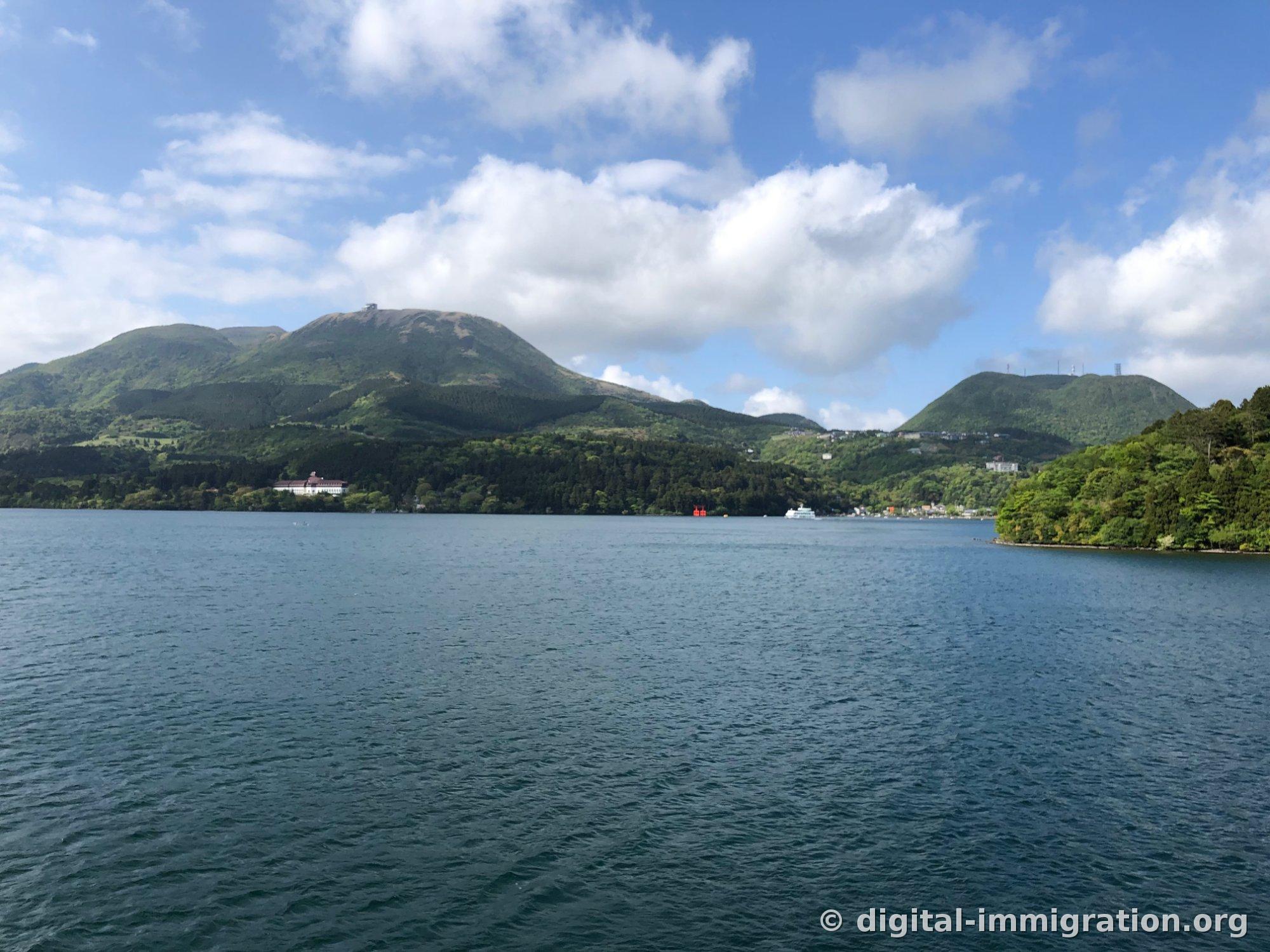 Surface bleu-vert profonde du lac Ashinoko avec des crêtes vertes s'étendant au-delà, et un petit torii rouge trempant ses pieds dans l'eau visible sur le rivage, avec des bâtiments blancs et un bateau amarré