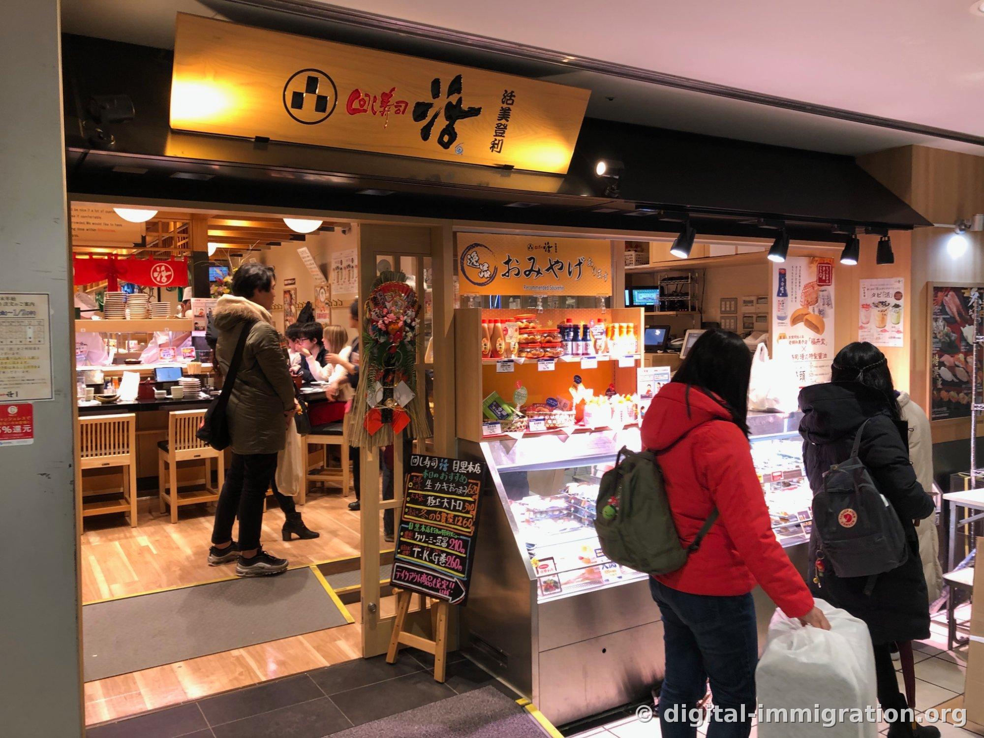 木质招牌上写着「回し寿司 活 活美登利」的卡茨美登利目黑本店入口，右边放着纪念品陈列柜和今日推荐菜单的黑板
