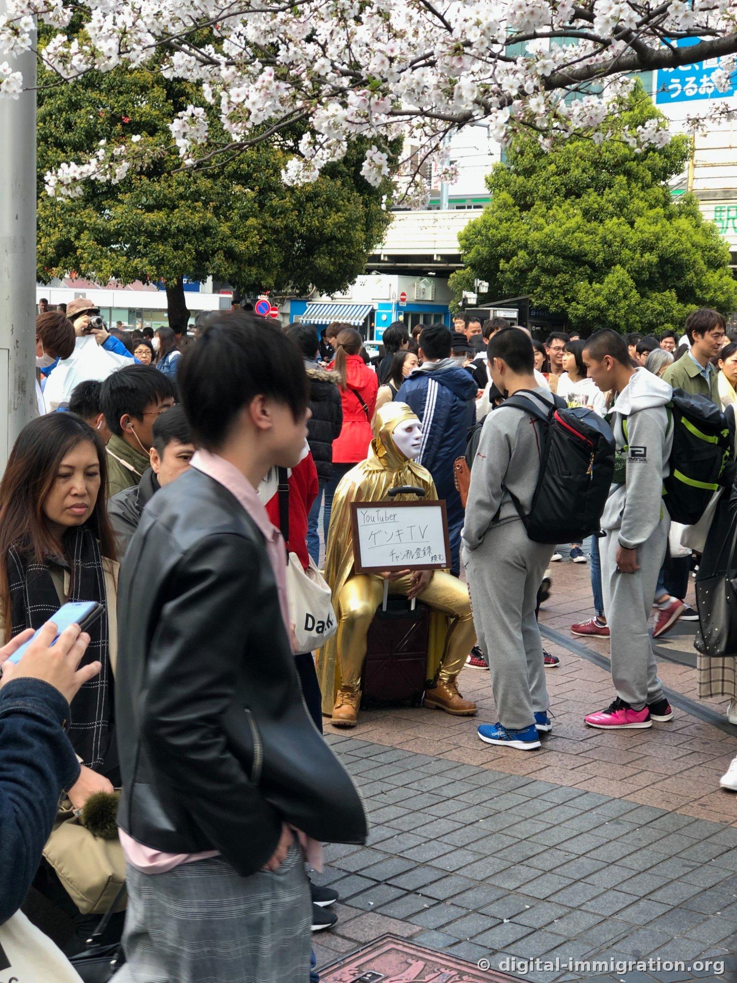 同じ金色の服のユーチューバーを縦に映した写真、周りはグレーのトレーニングウェアの人たちと人混みでいっぱい