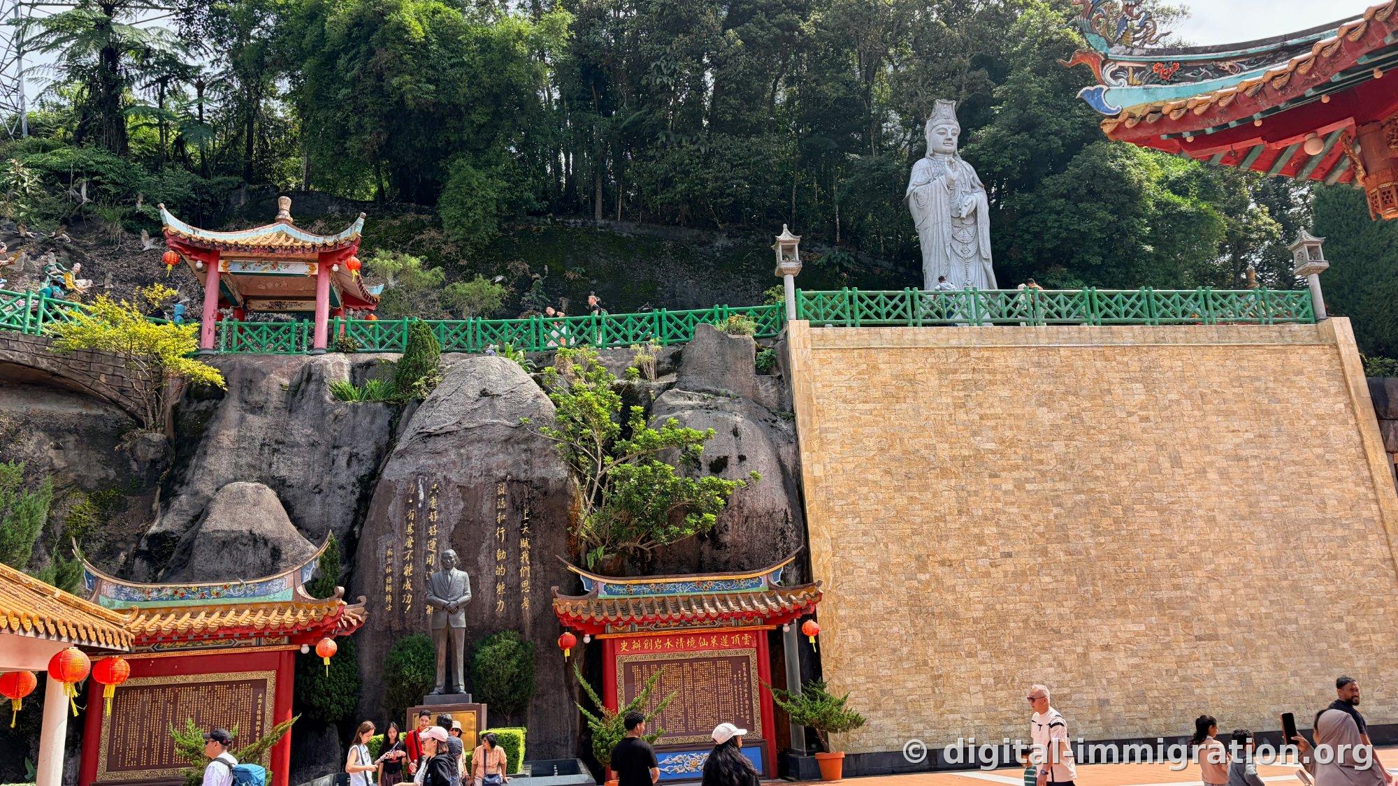 Une statue debout blanche de Guanyin derrière une balustrade verte, un pavillon rouge au-dessus, avec un mur de falaise rocheuse, une statue en costume d'homme et une roche gravée au-dessous