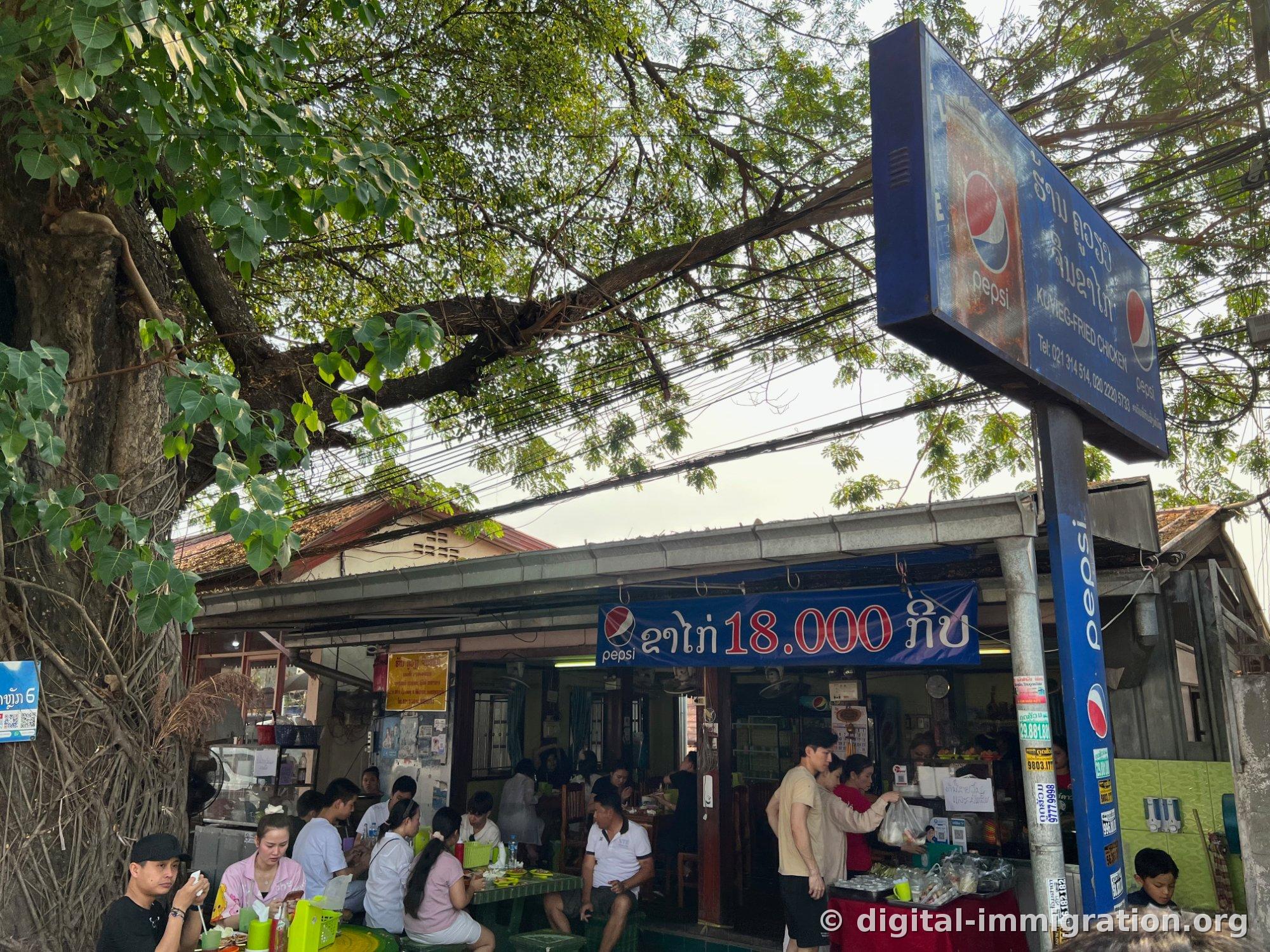 A Lao fried chicken shop under a large tree with a blue Pepsi sign and 'KO-VIENG FRIED CHICKEN' written on it, with people sitting at outdoor tables