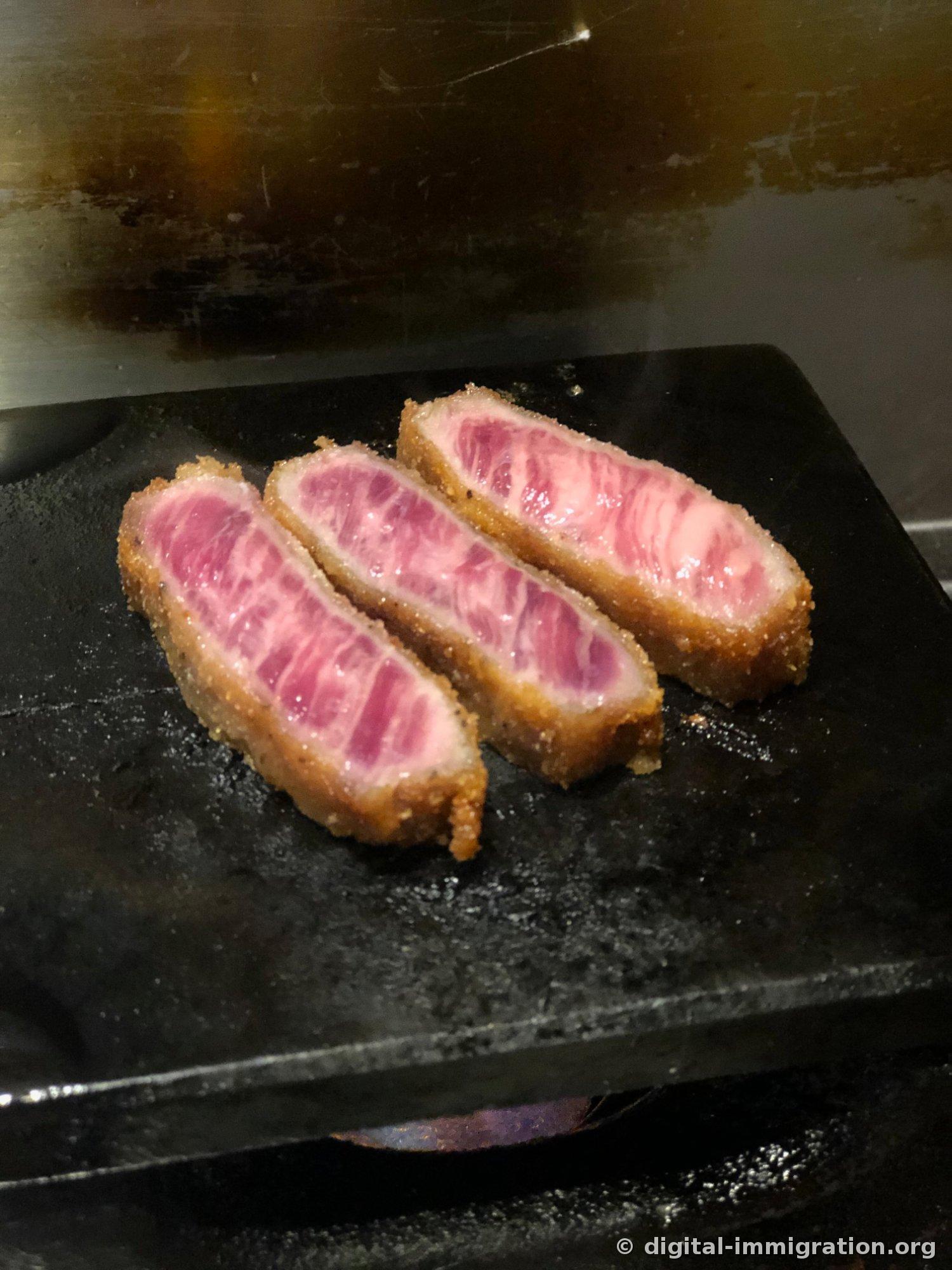 Trois morceaux de Gyukatsu juste posés sur la pierre noire. L'intérieur de la tranche est encore un rose vif de viande crue avec du marbling visible
