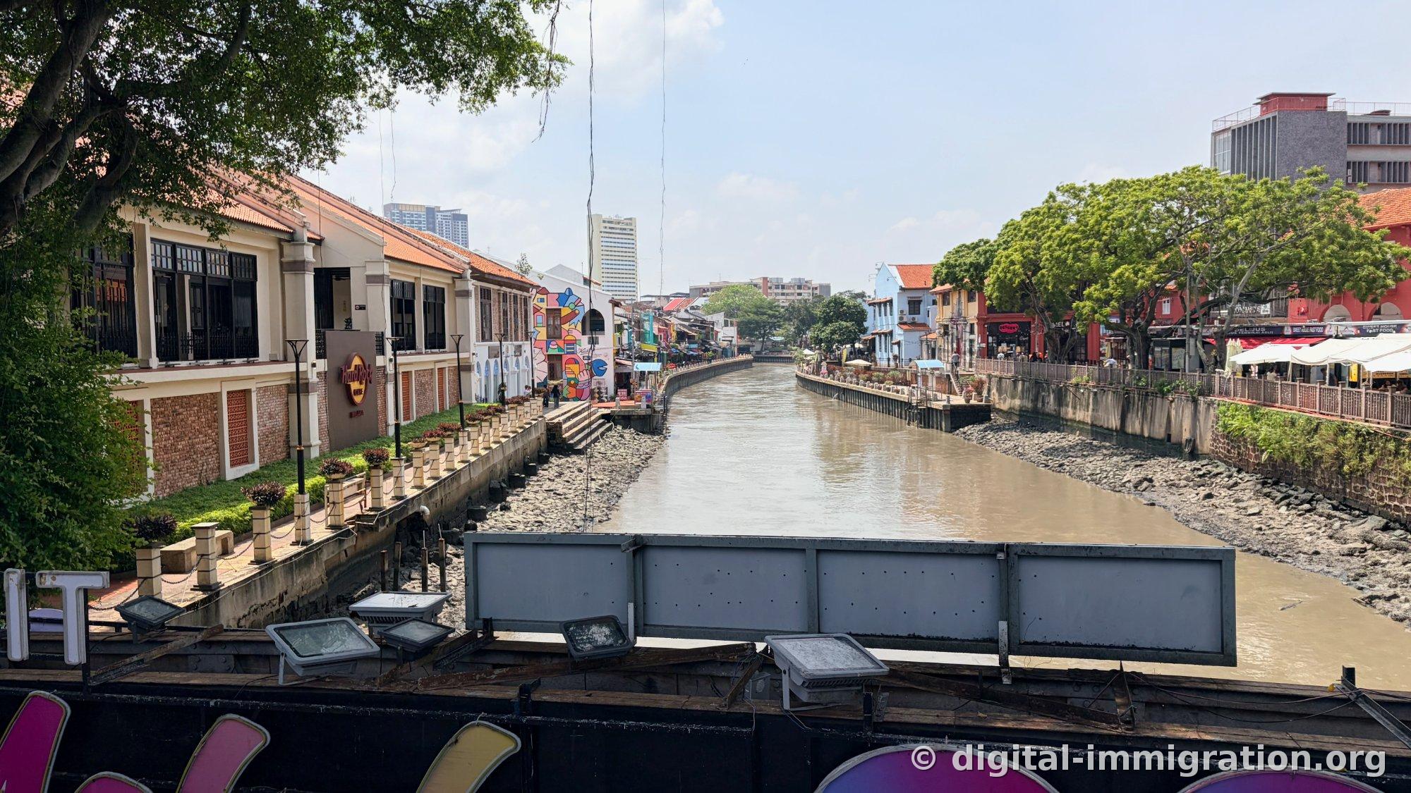 The Melaka River as seen from the Tan Kim Seng bridge. A murky river flows between a Hard Rock Cafe and shophouses with murals on the left, and open-air restaurants with parasols on the right