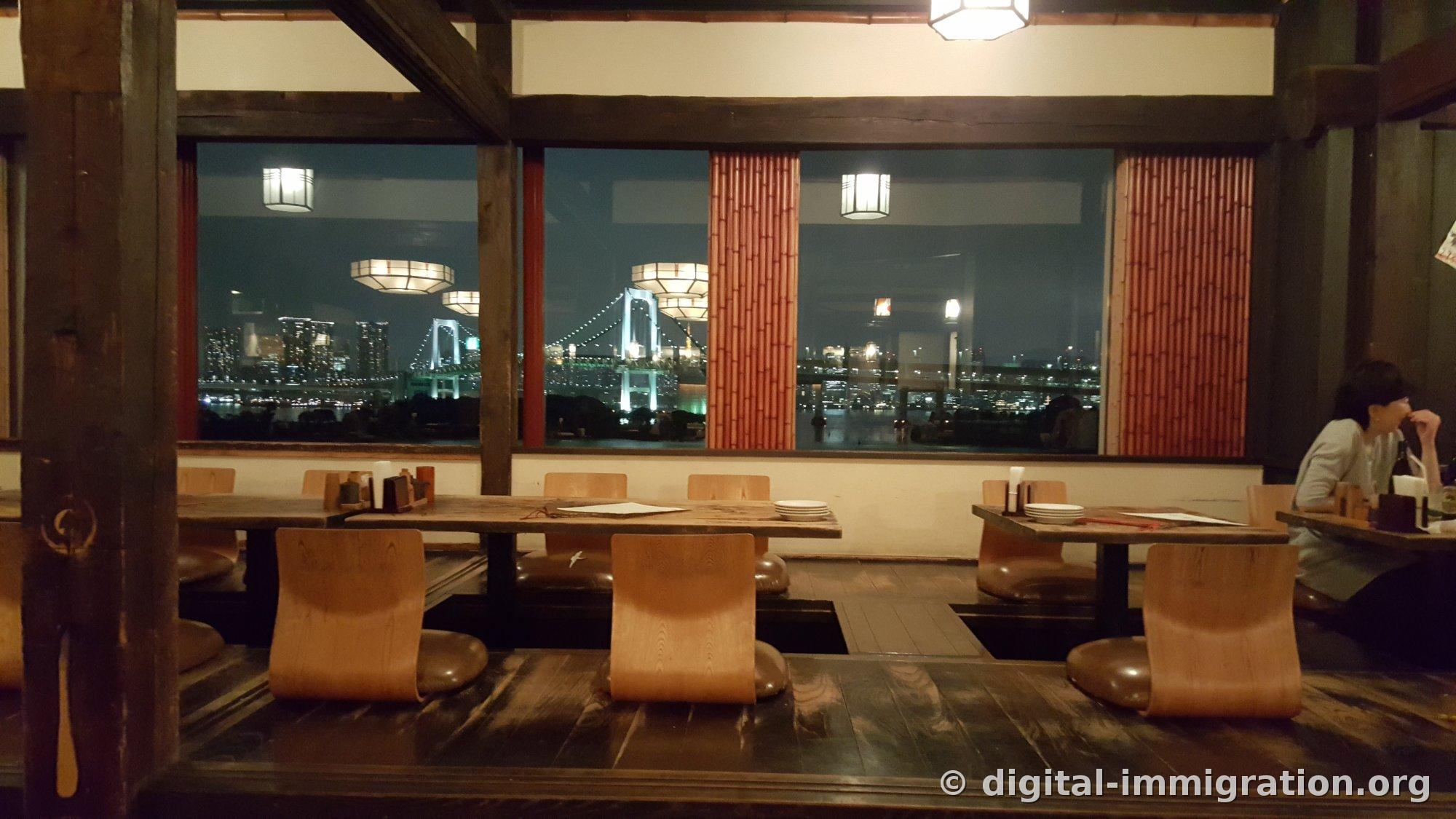 A view from inside a dimly lit wooden interior seating restaurant, with a large window framing the Rainbow Bridge glowing in blue and Tokyo's nightscape beyond, indoor lighting reflected on the glass overlapping with the bridge