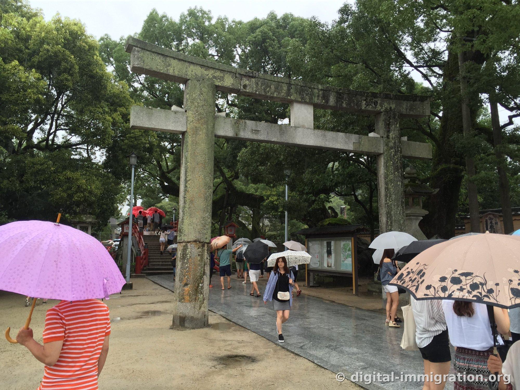 Worshippers with umbrellas passing under a large moss-covered stone torii at the entrance to Dazaifu Tenmangu on a rainy day