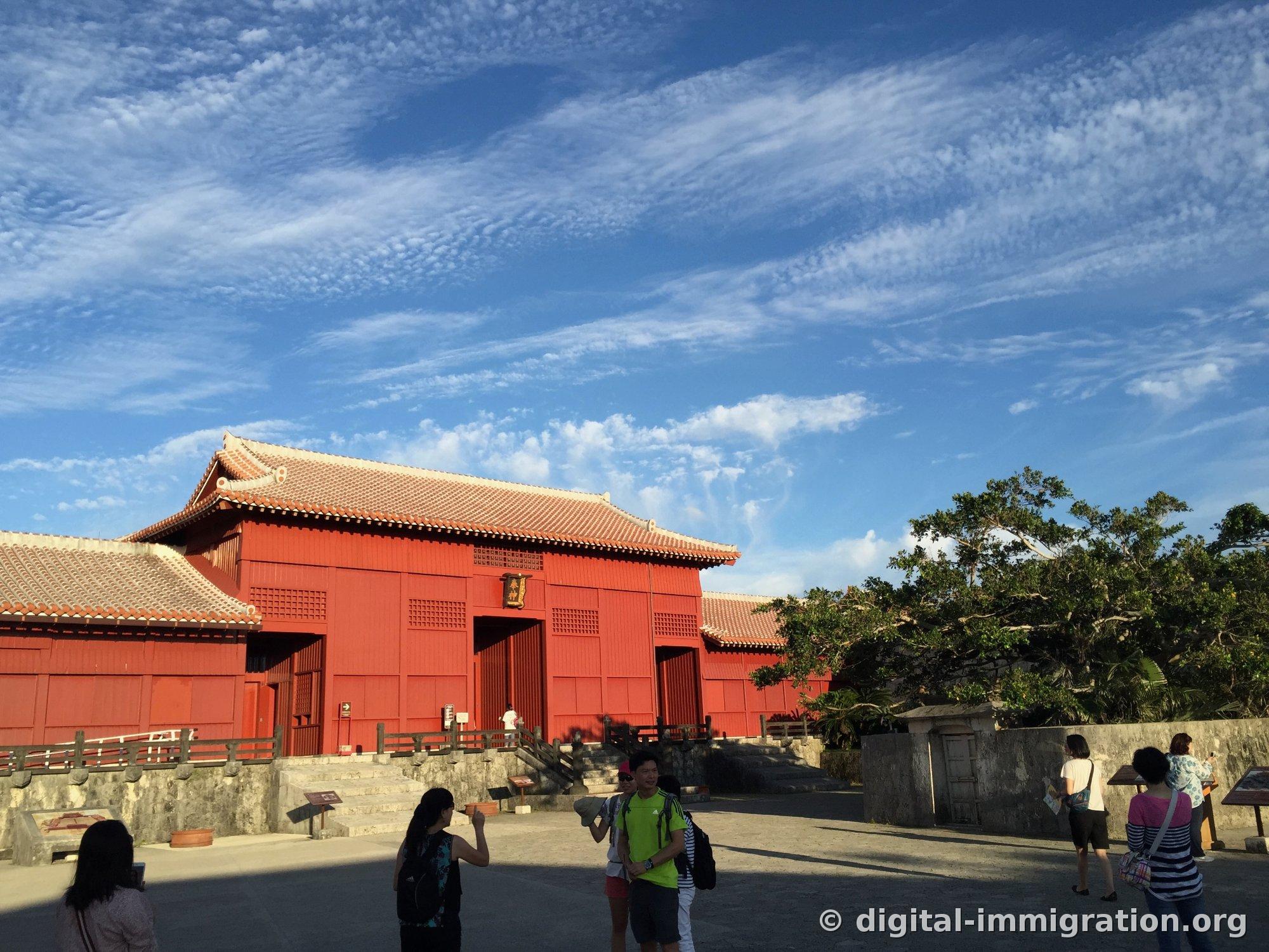 Houshinmon with vermillion walls and red-tiled roof, scattered clouds against a wide sky, tourists in the plaza below