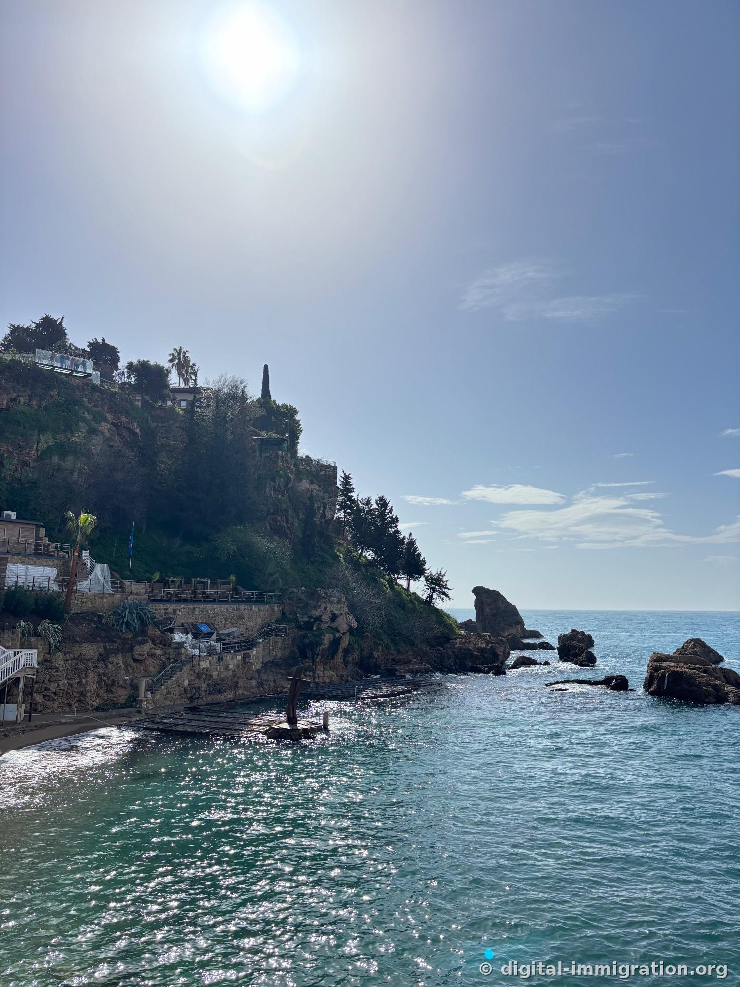 Backlit cliff-side beach with small cove, floating dock half-installed, sunbeds wrapped in white cloth, glass viewing platform with people standing on cliff edge above