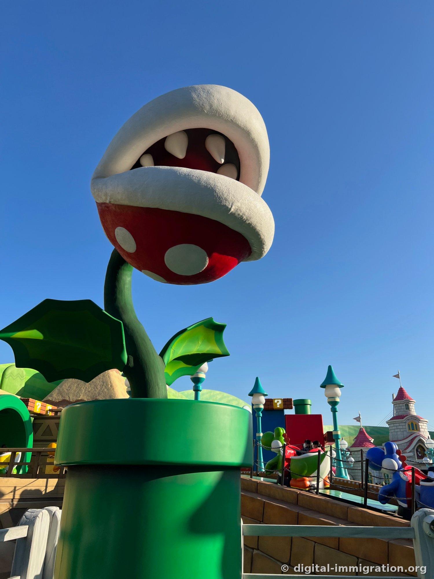 A giant Piranha Plant sculpture emerging from a green pipe against a blue sky, with Peach's Castle spire visible in the background