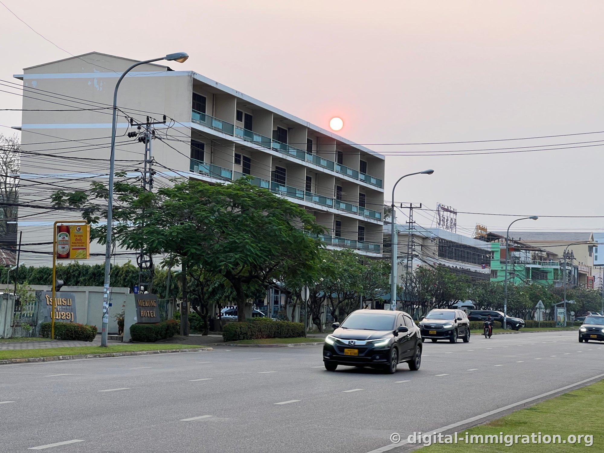 夕方のビエンチャン幹線道路、MEKONG HOTELの看板がある4階建ての建物と絡み合う電線の電柱、通り過ぎる黒いホンダの車両