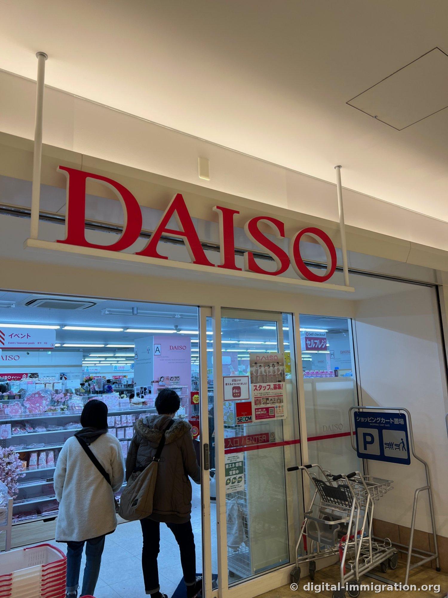 L'entrée d'un magasin DAISO avec son enseigne rouge, des chariots de shopping, et deux clients entrant